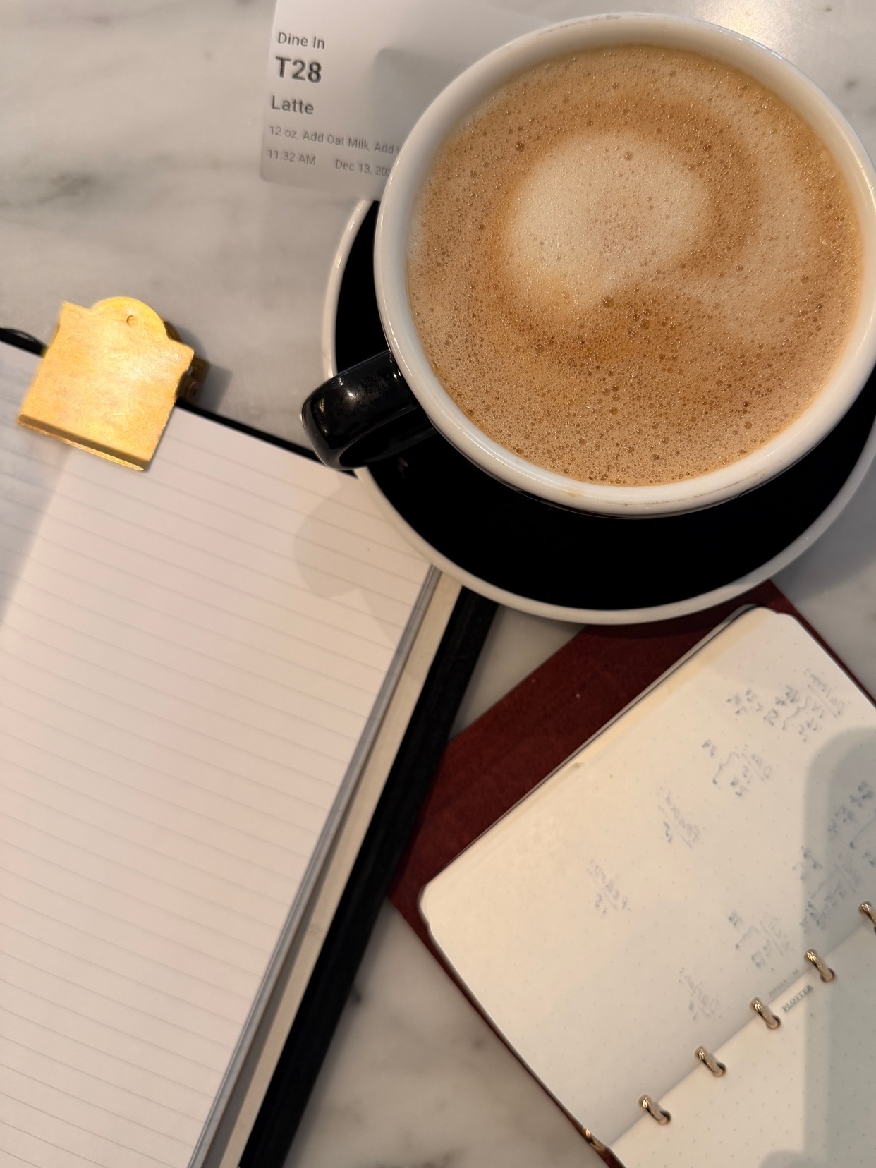 A close-up shot of a latte in a black mug, sitting on a saucer, next to open notebooks and a gold paper clip. The scene is set on a marble table.