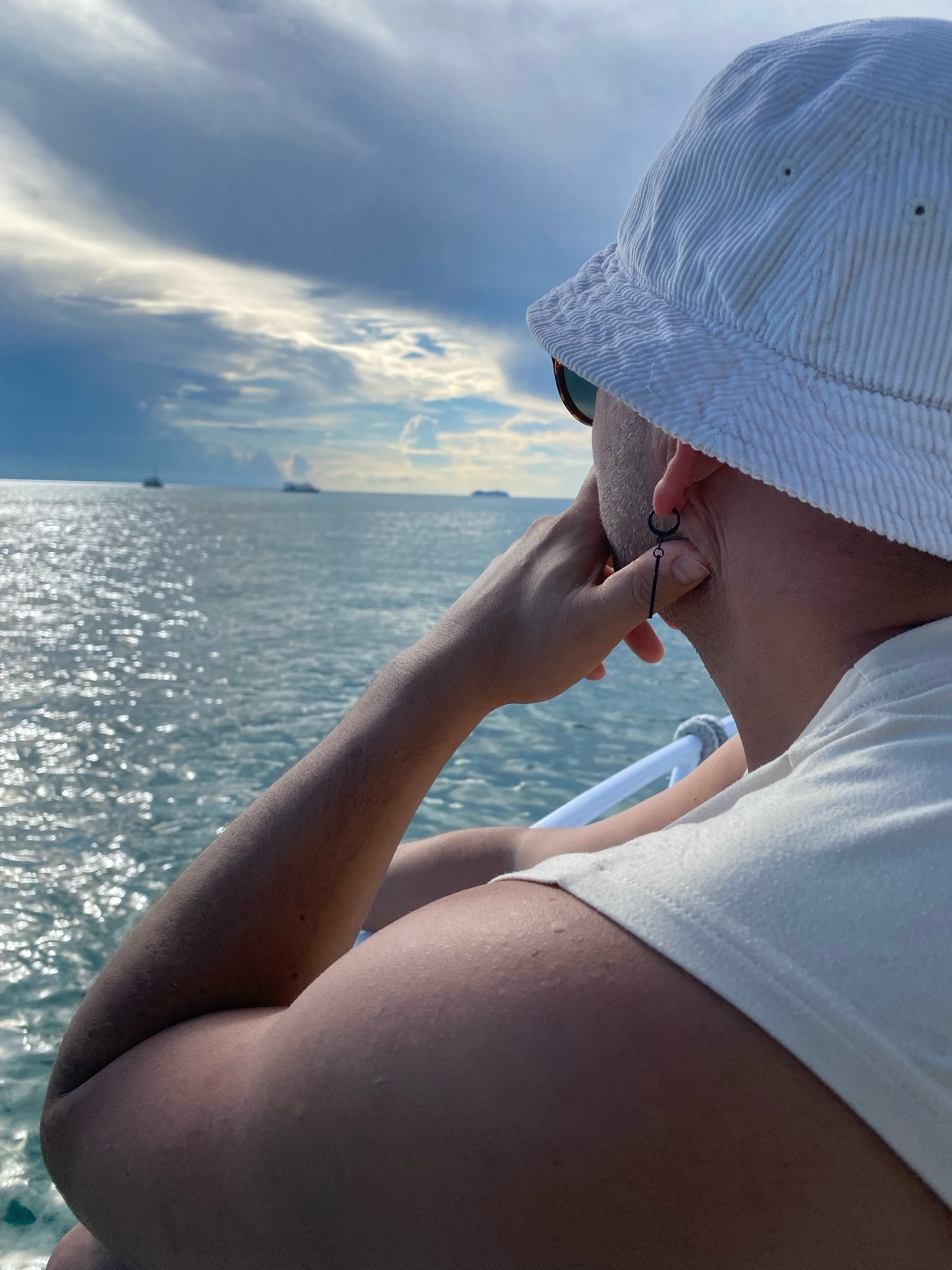 A man wearing a bucket hat and sunglasses looks out at the ocean. The sky is cloudy, and the water is calm.