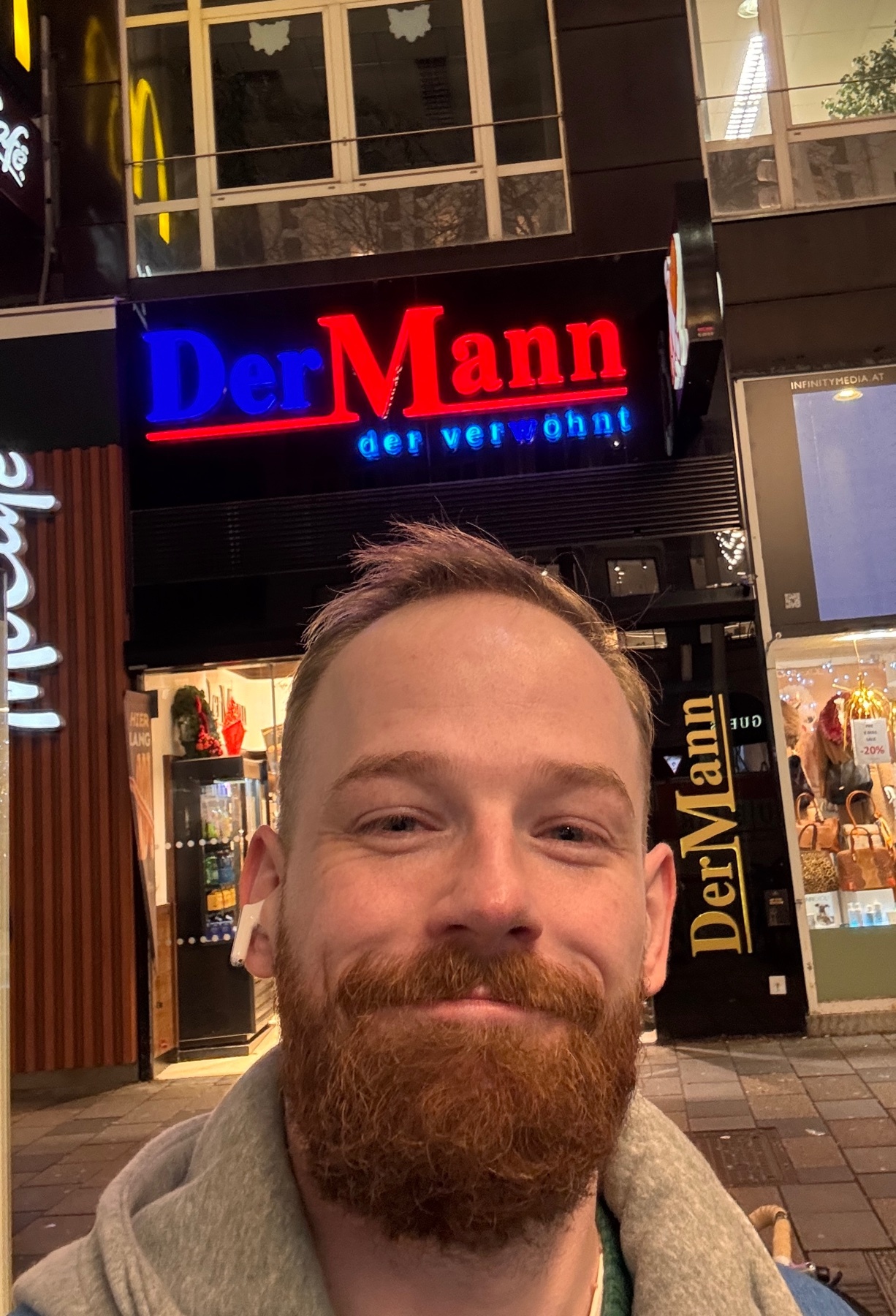A man takes a selfie in front of a shop with German signage. The shop is lit up at night.