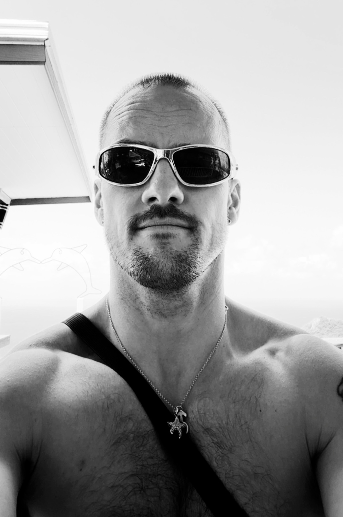 A black and white, close-up selfie of a man wearing sunglasses and a necklace. He has stubble and a bald head.