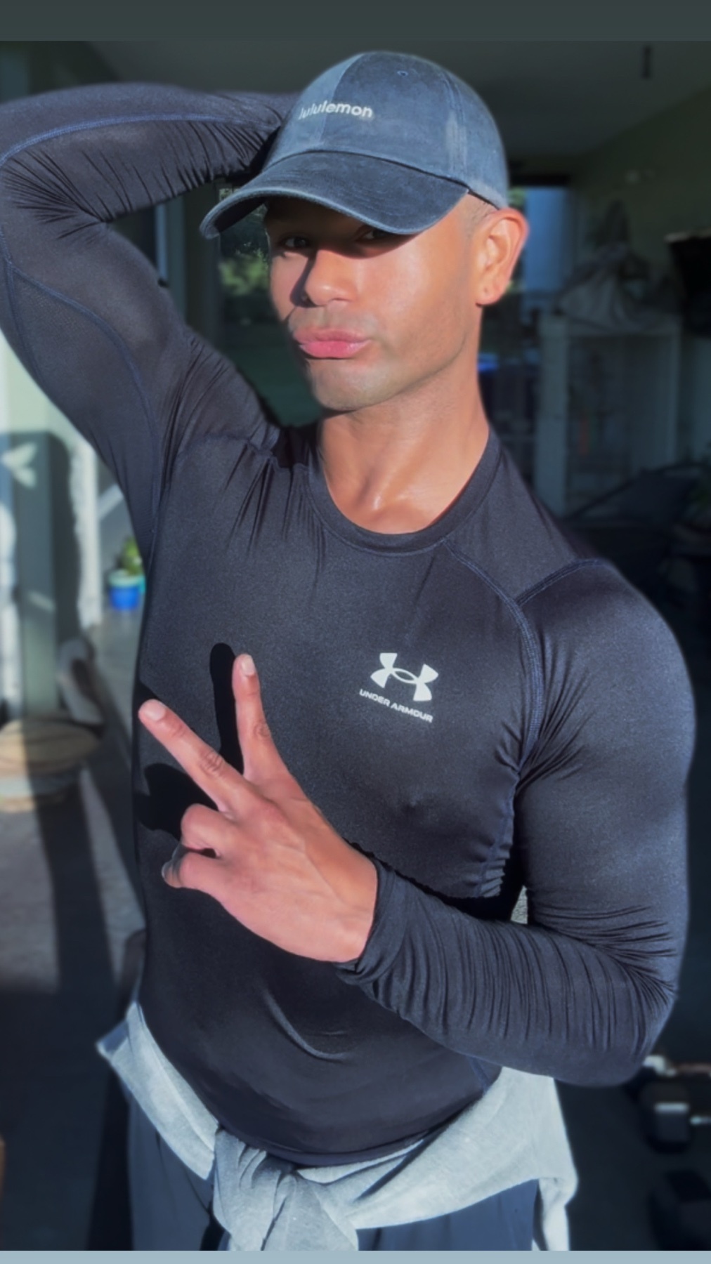 A man poses for a selfie in a gym, flexing his arm. He is wearing a long-sleeved shirt and a cap.