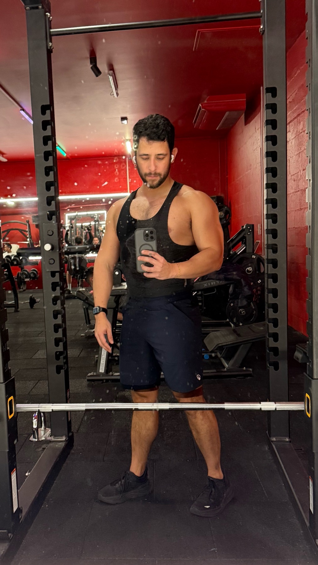 A man takes a mirror selfie at the gym. He is wearing a tank top and shorts.