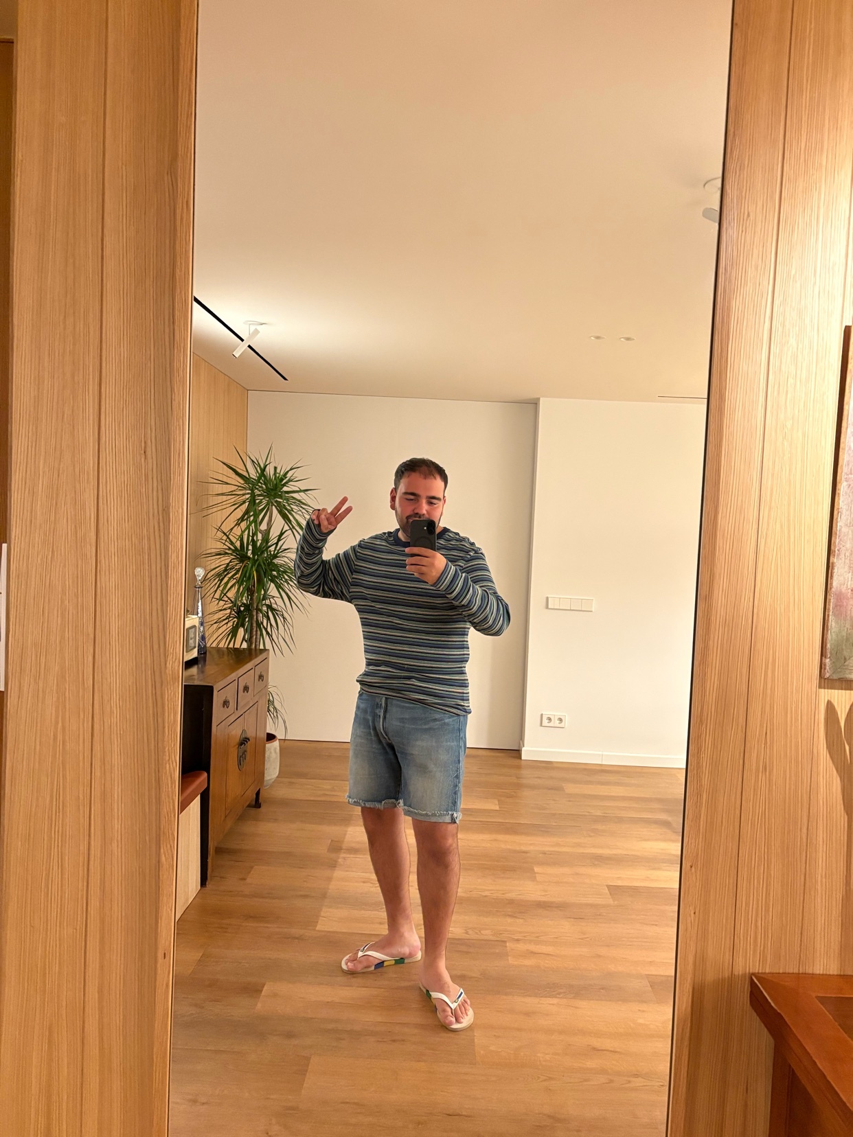 A man takes a mirror selfie while wearing a striped long-sleeved shirt, denim shorts, and flip-flops. He is standing in a room with wooden floors and furniture, holding up two fingers in a peace sign.