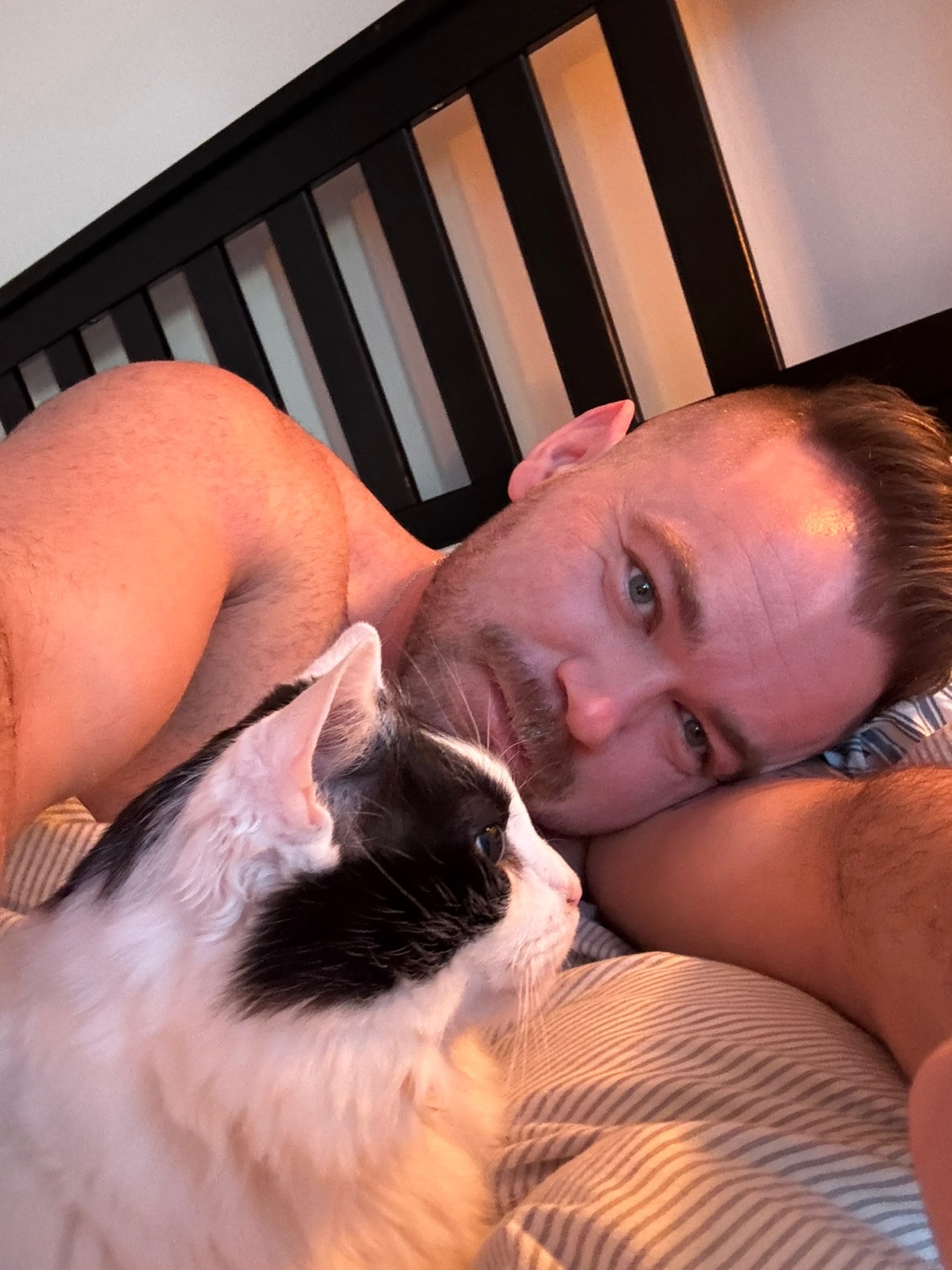 A man with short hair and stubble lies in bed with a black and white cat. The man is looking towards the camera while the cat looks away.