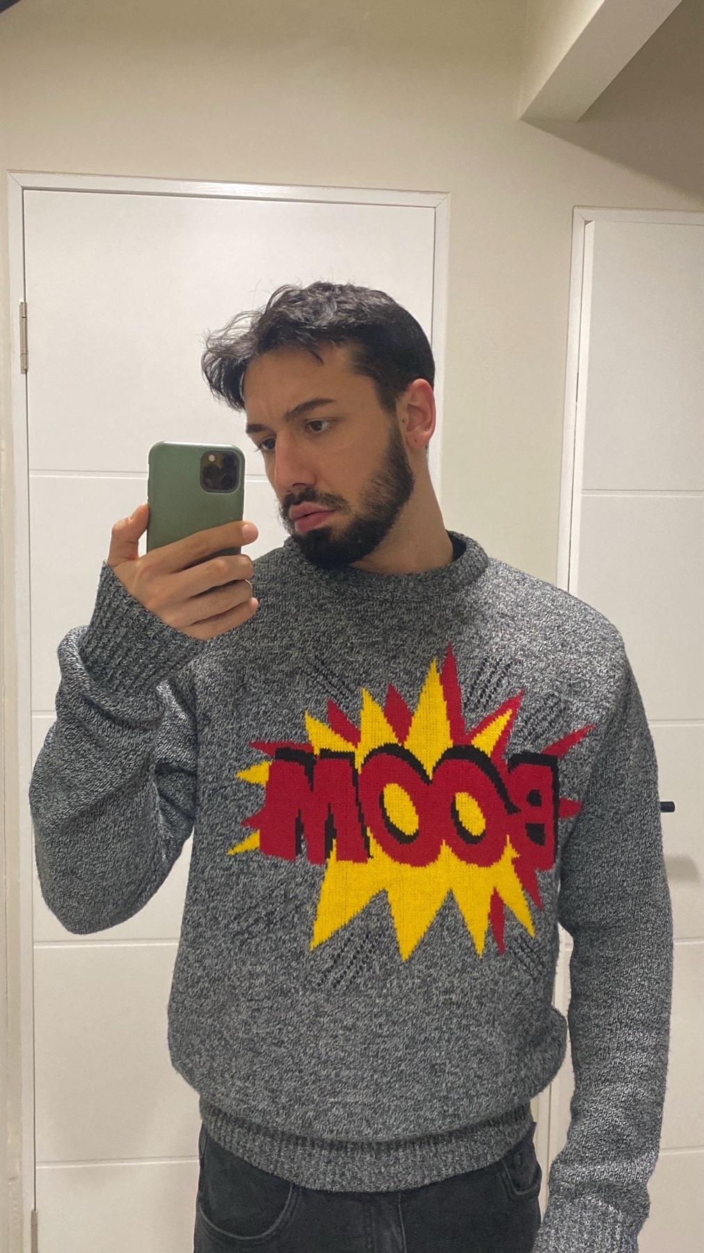 A man takes a mirror selfie, wearing a gray sweater with a "BOOM" graphic. The image is a modern style.