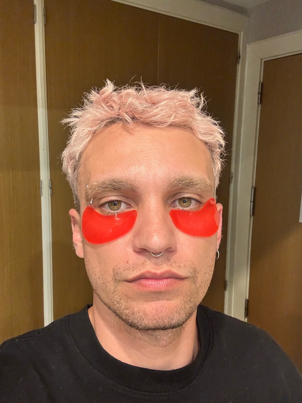 A man with pink hair and a nose ring poses for a selfie. He is wearing a black t-shirt and red eye masks.