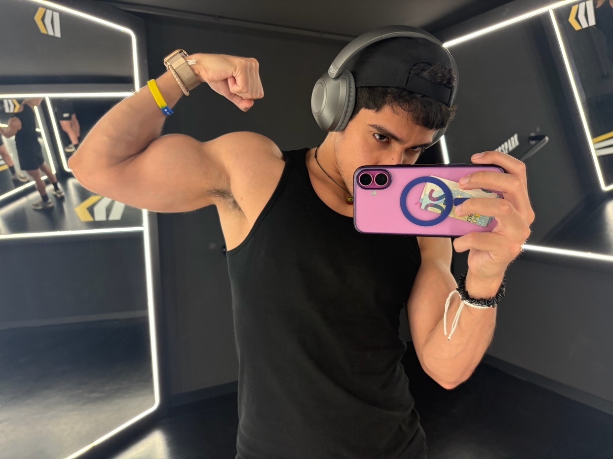 A man in a black tank top and headphones flexes his bicep in a gym selfie. The man is looking at his phone as he takes the picture, with mirrors reflecting the gym environment behind him.