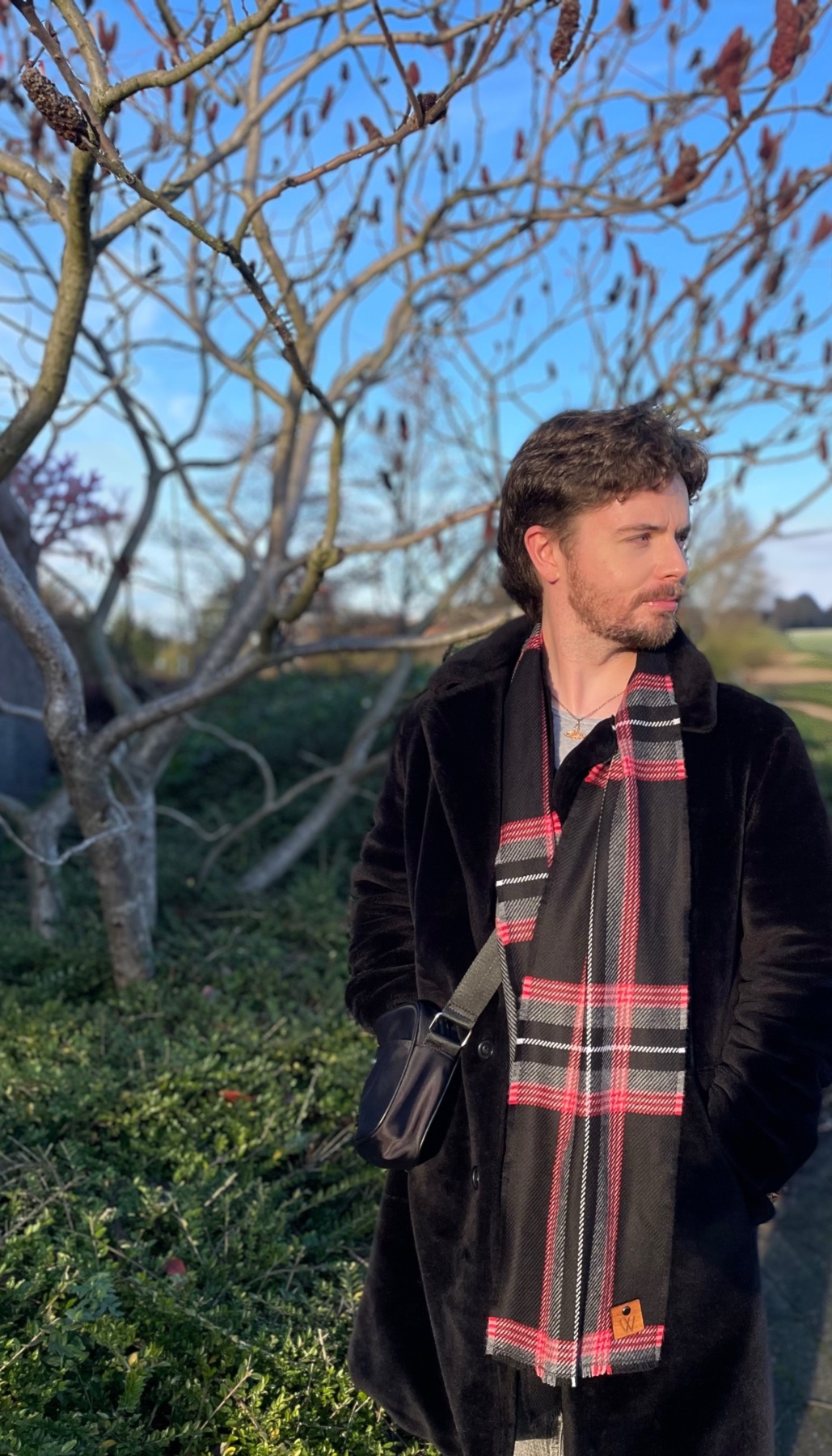 A man poses outdoors, wearing a stylish coat and scarf. The photo is taken during the day, with a tree in the background.