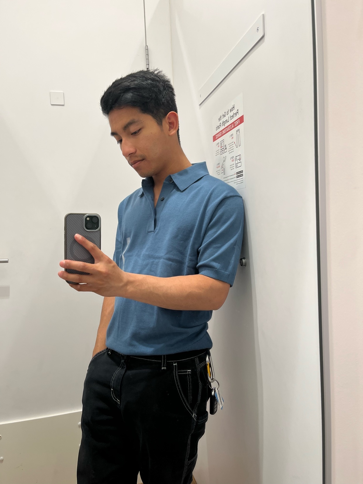 A man takes a selfie in a fitting room, wearing a blue polo shirt and black pants.