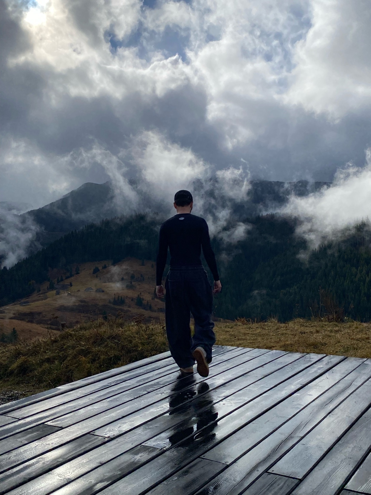A man stands on a wooden platform, gazing at a mountain range shrouded in clouds. The photo captures a moment of solitude and appreciation for nature.