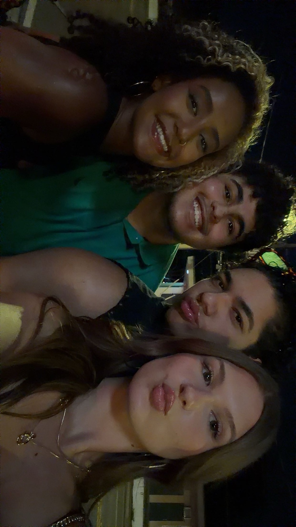 A group of four friends smile and pose for a selfie at night. The two women in the foreground pout their lips, while the man and woman behind them beam at the camera.
