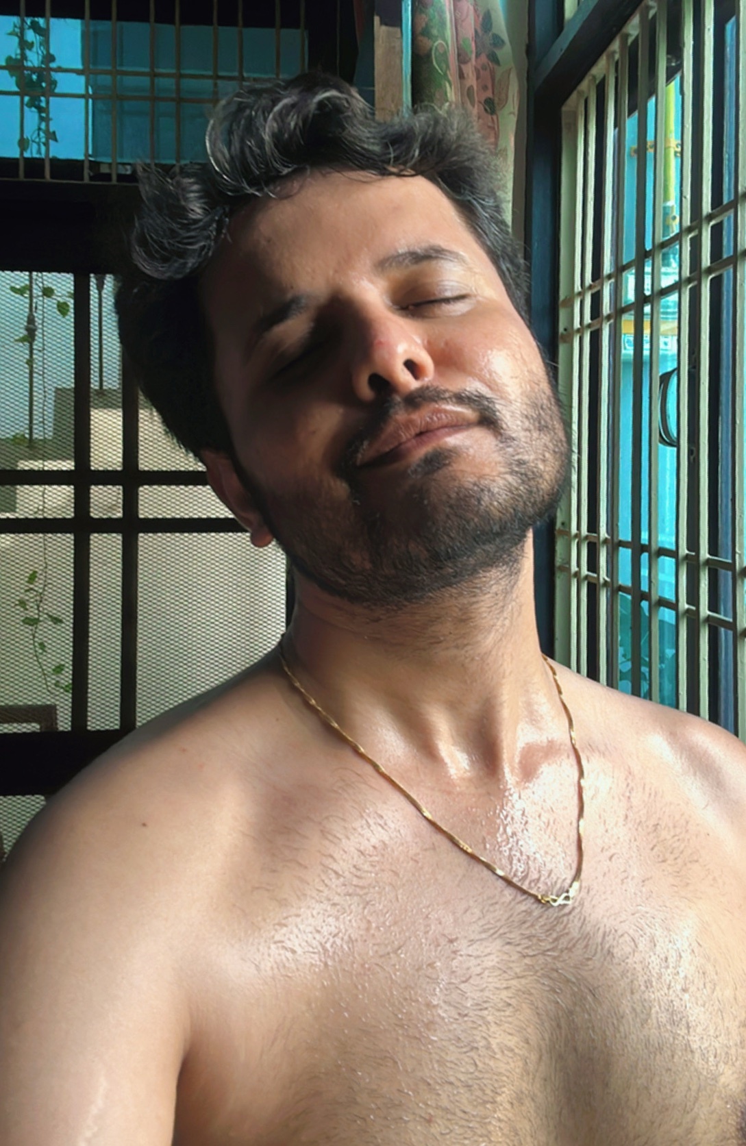 A shirtless man with dark hair and a beard is sweating. He is wearing a gold necklace and appears to be indoors.