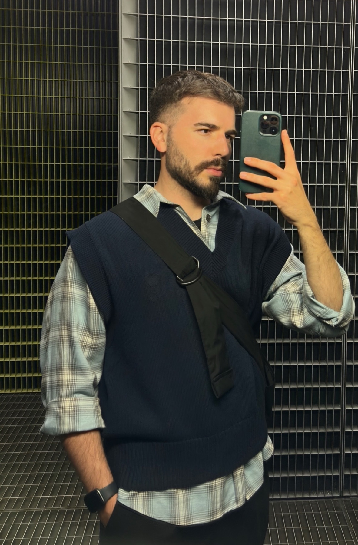 A man takes a mirror selfie. He is wearing a plaid shirt, a dark blue sweater vest, and a black bag.