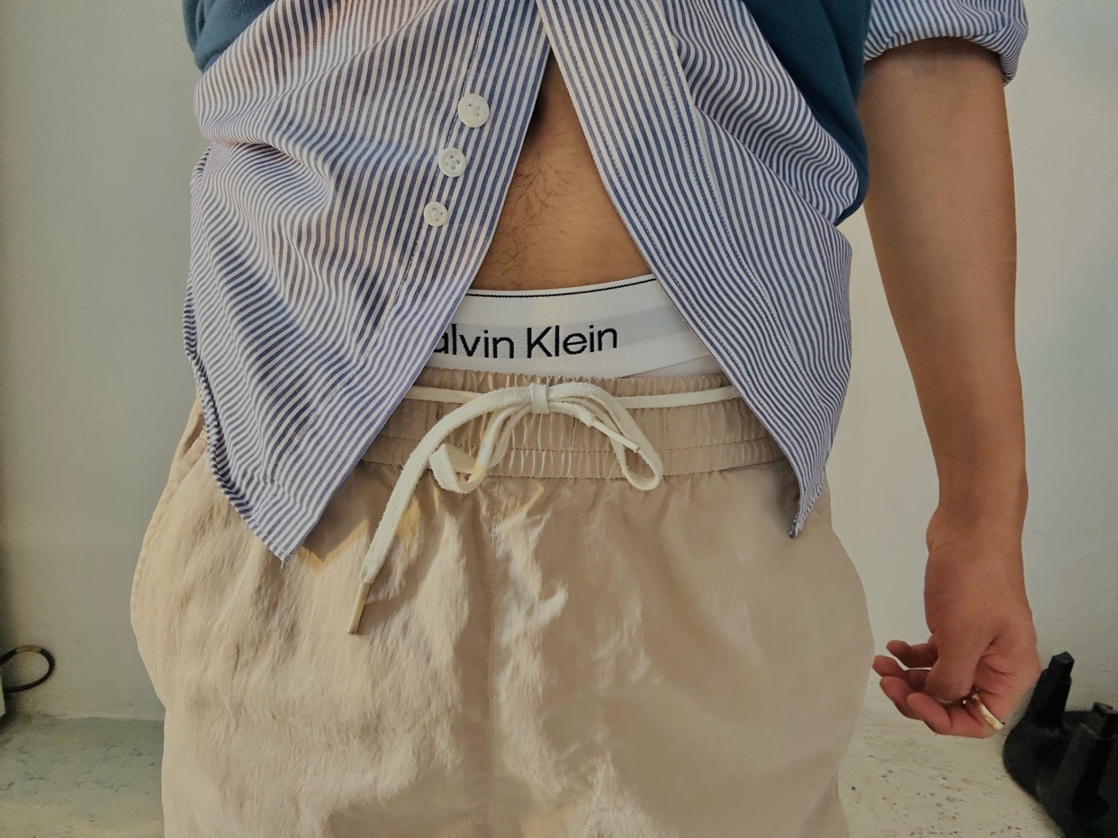 A close-up shot focuses on a man's lower torso, showcasing his Calvin Klein branded underwear peeking out from under his light-colored drawstring shorts. He is wearing a blue and white striped button-down shirt partially unbuttoned, revealing his mid