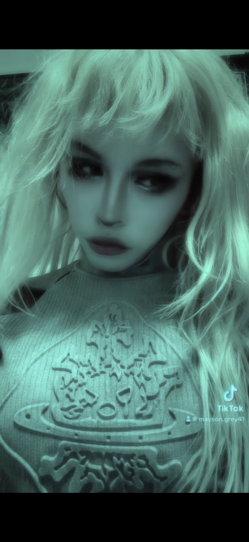 A woman with blonde hair and doll-like features poses for a selfie. The image has an alternative aesthetic.