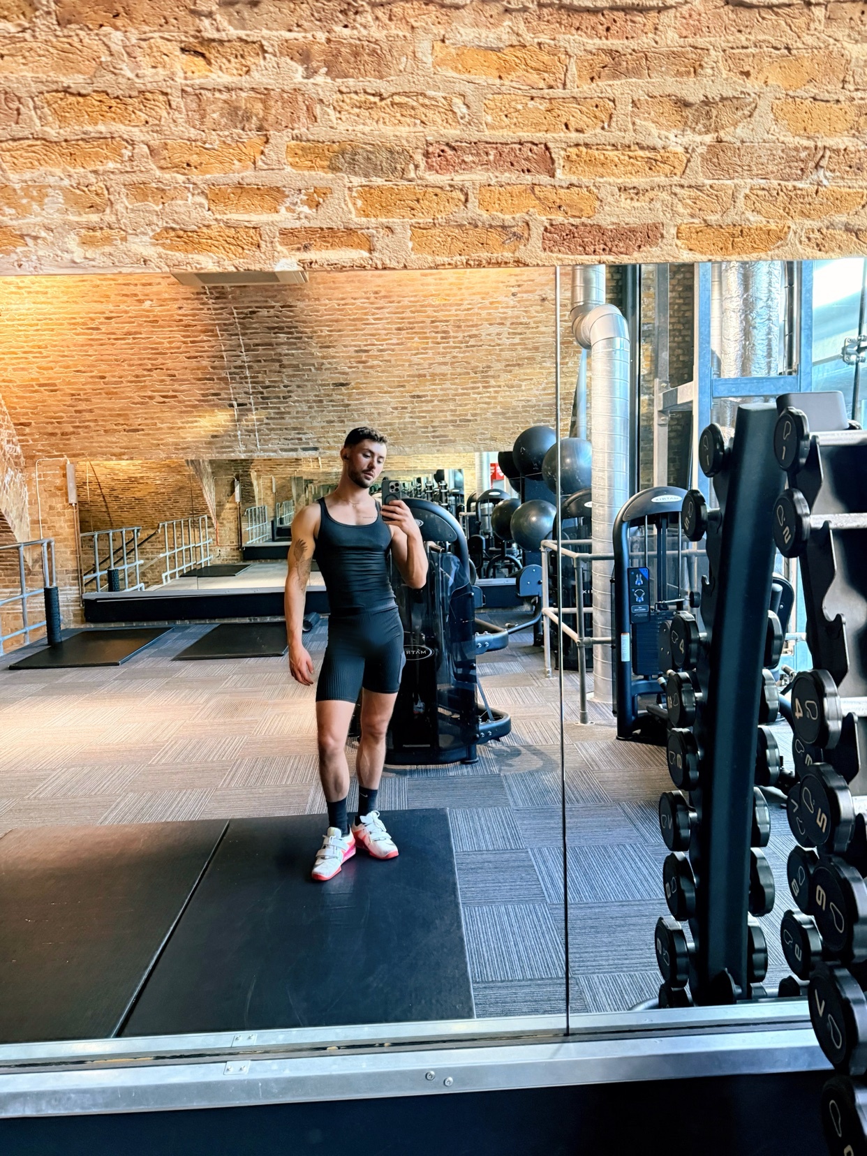 A man takes a mirror selfie in a gym. He is wearing a black tank top and shorts, showcasing his muscular physique.