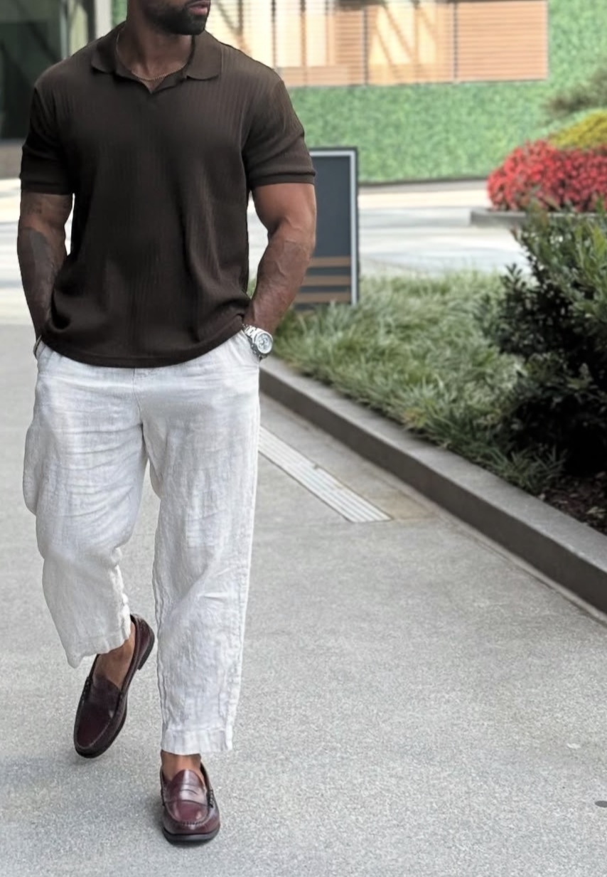 A man in a stylish outfit walks down a sidewalk. He wears a brown polo shirt, white linen pants, and brown loafers.