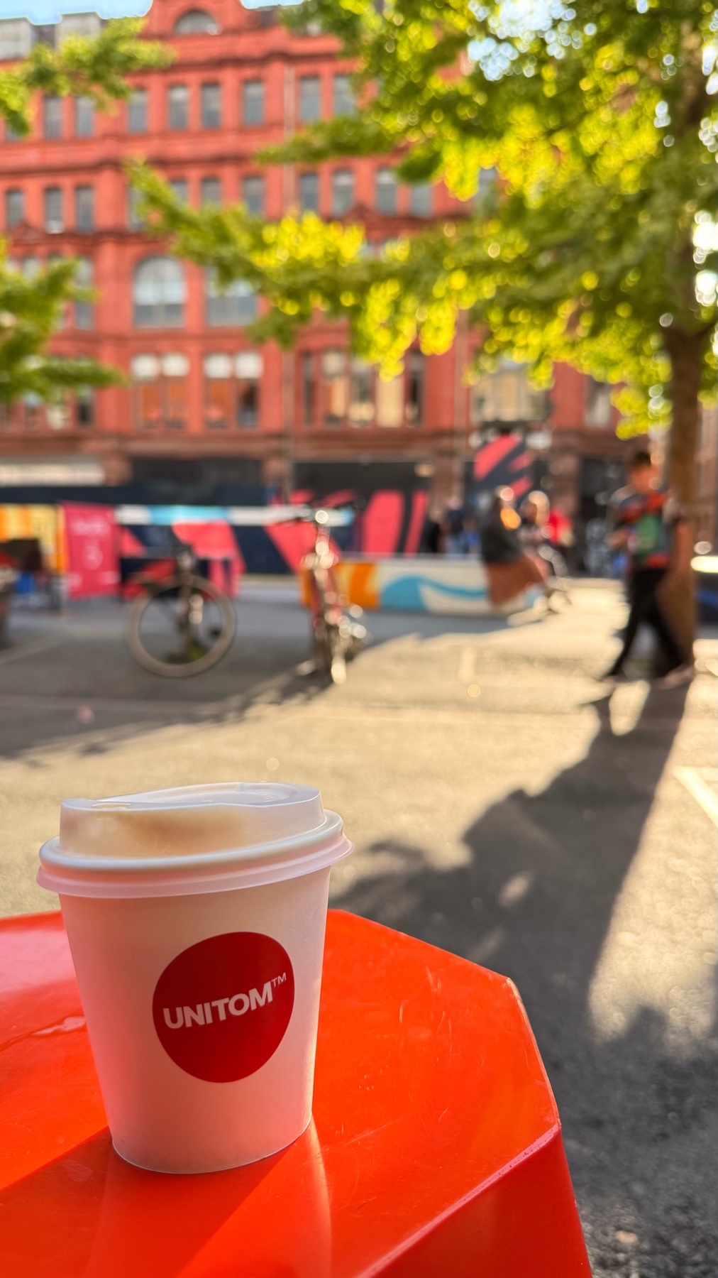A white paper coffee cup with a red logo sits on an orange table in the foreground. In the blurred background, a red brick building, trees, and people are visible on a sunny day.