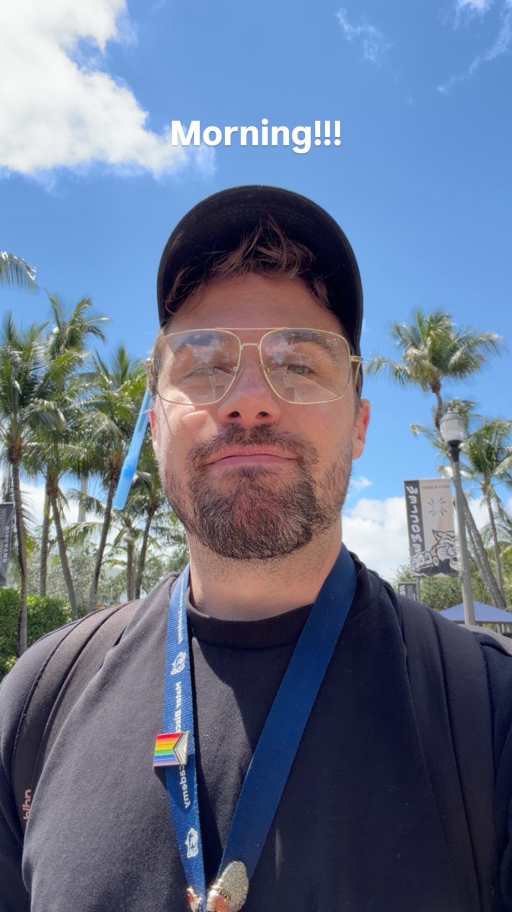 A man with glasses and a beard takes a selfie outdoors with palm trees and a blue sky in the background. He is wearing a black t-shirt and a baseball cap.