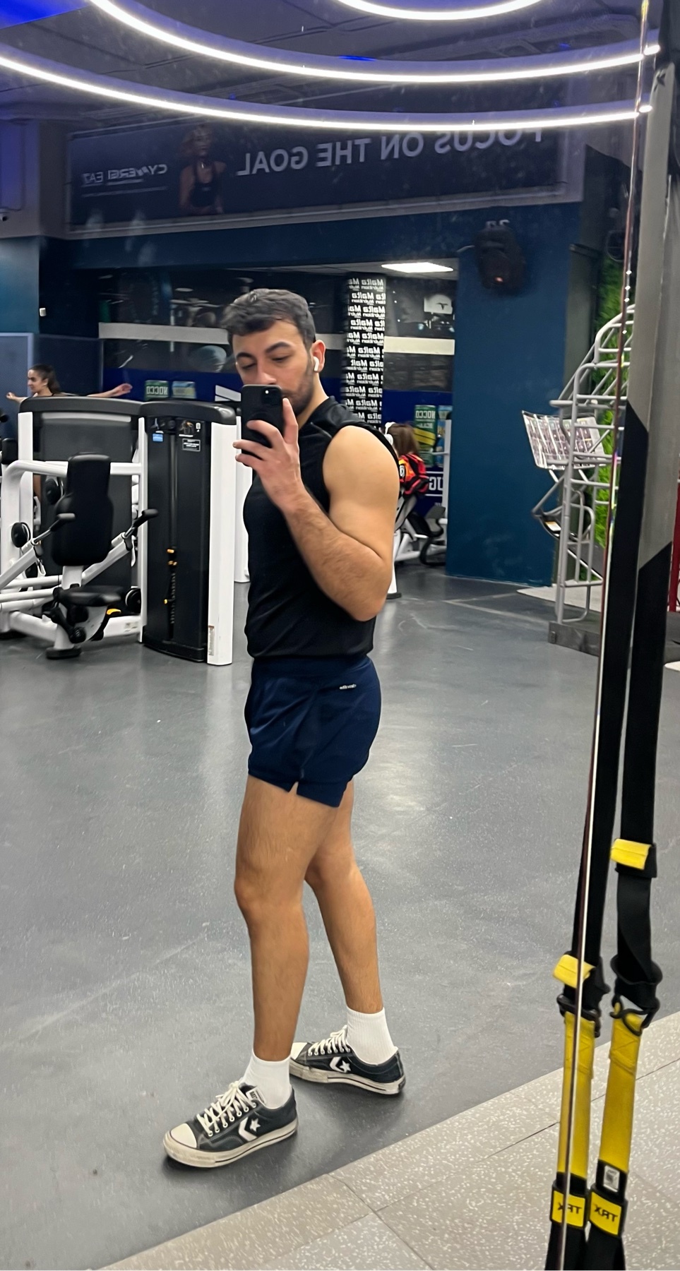 A man takes a mirror selfie at the gym, flexing his arm. He is wearing shorts and sneakers.