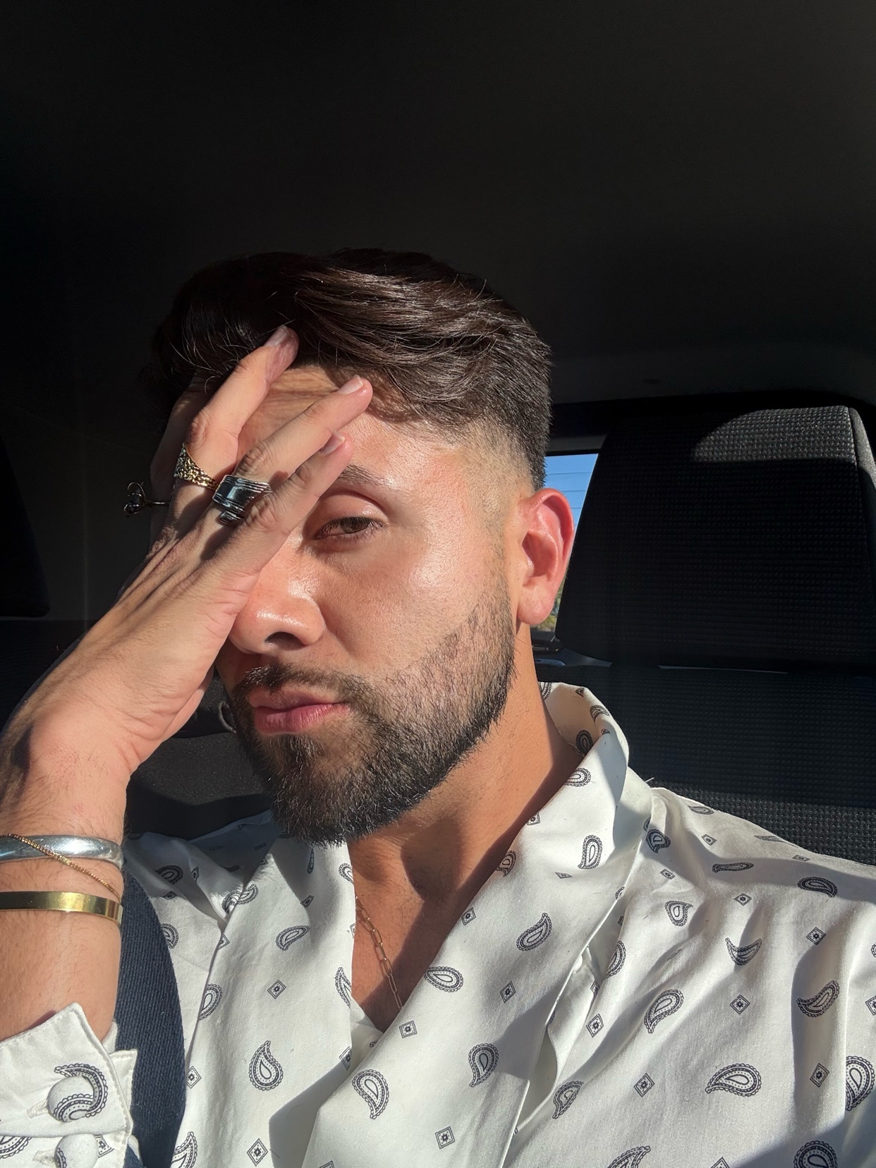 A man with a beard and dark hair poses for a selfie, shielding his eyes from the sun with his hand. He wears a patterned shirt and several rings and bracelets.