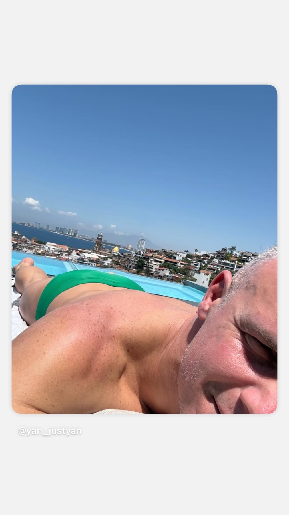 A man in green swimming trunks lies poolside with a cityscape and ocean in the background under a clear blue sky.