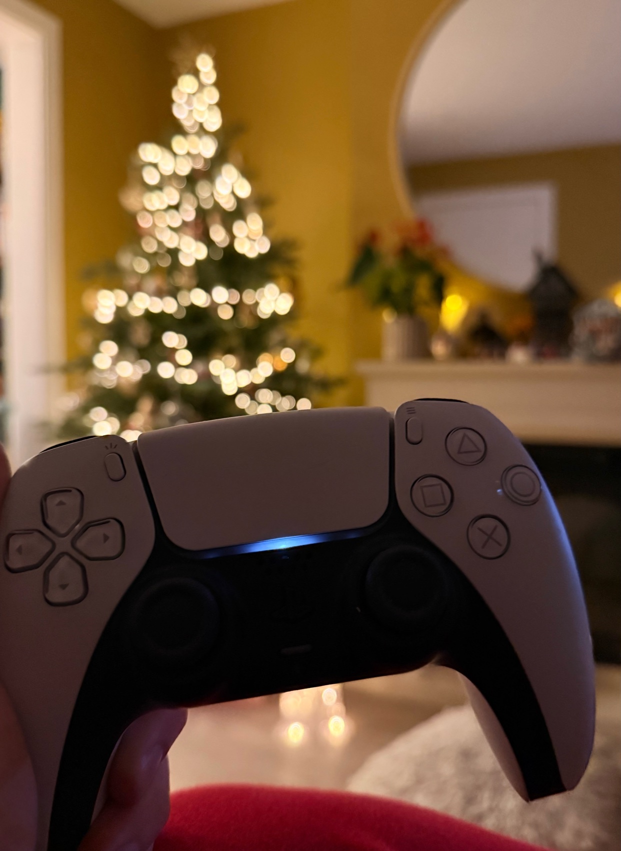 A person holding a Playstation 5 controller with a Christmas tree in the background. The image captures a cozy gaming setup during the holiday season.