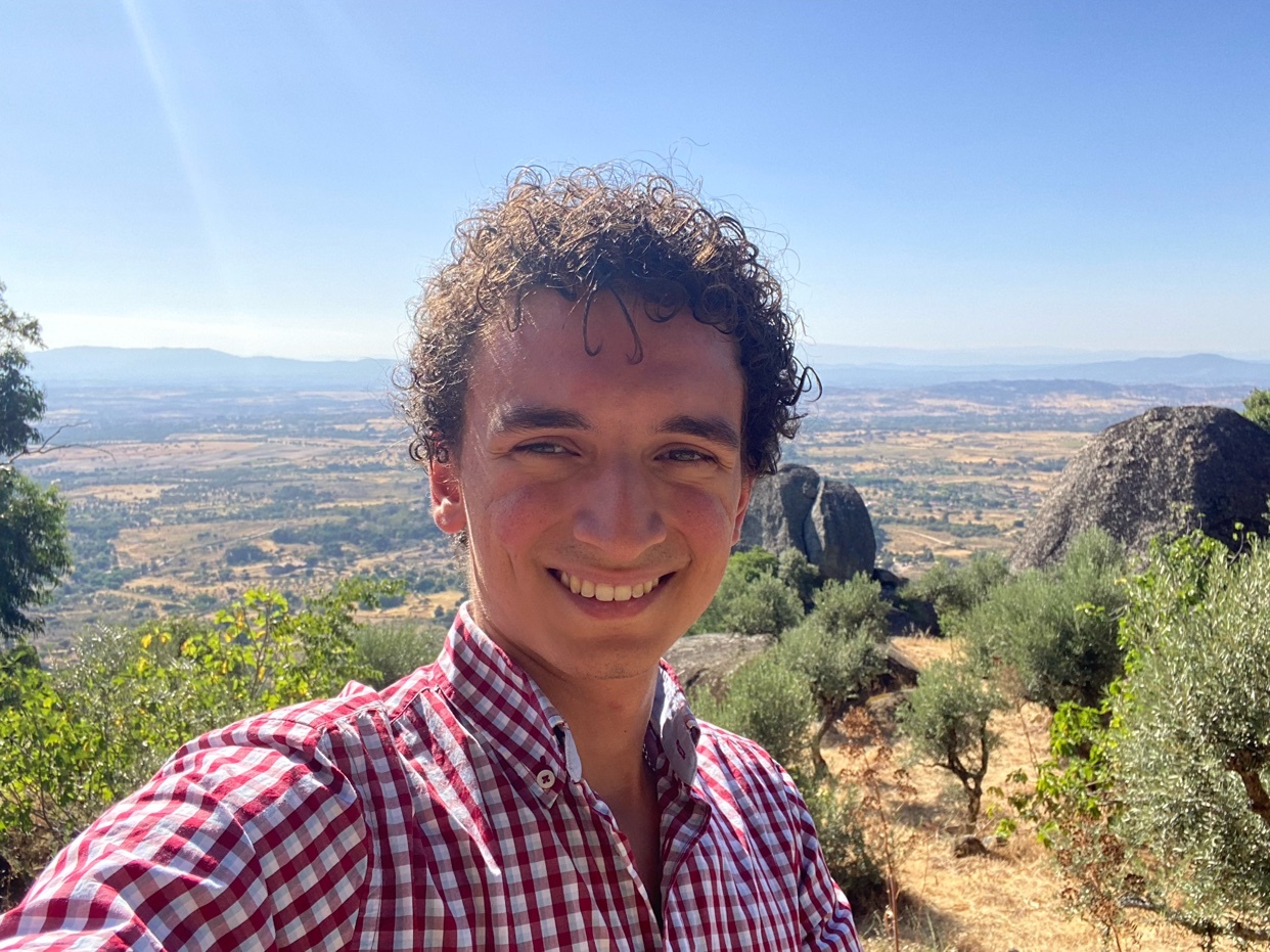 A young man with curly hair smiles at the camera in a selfie taken outdoors with a scenic landscape in the background.