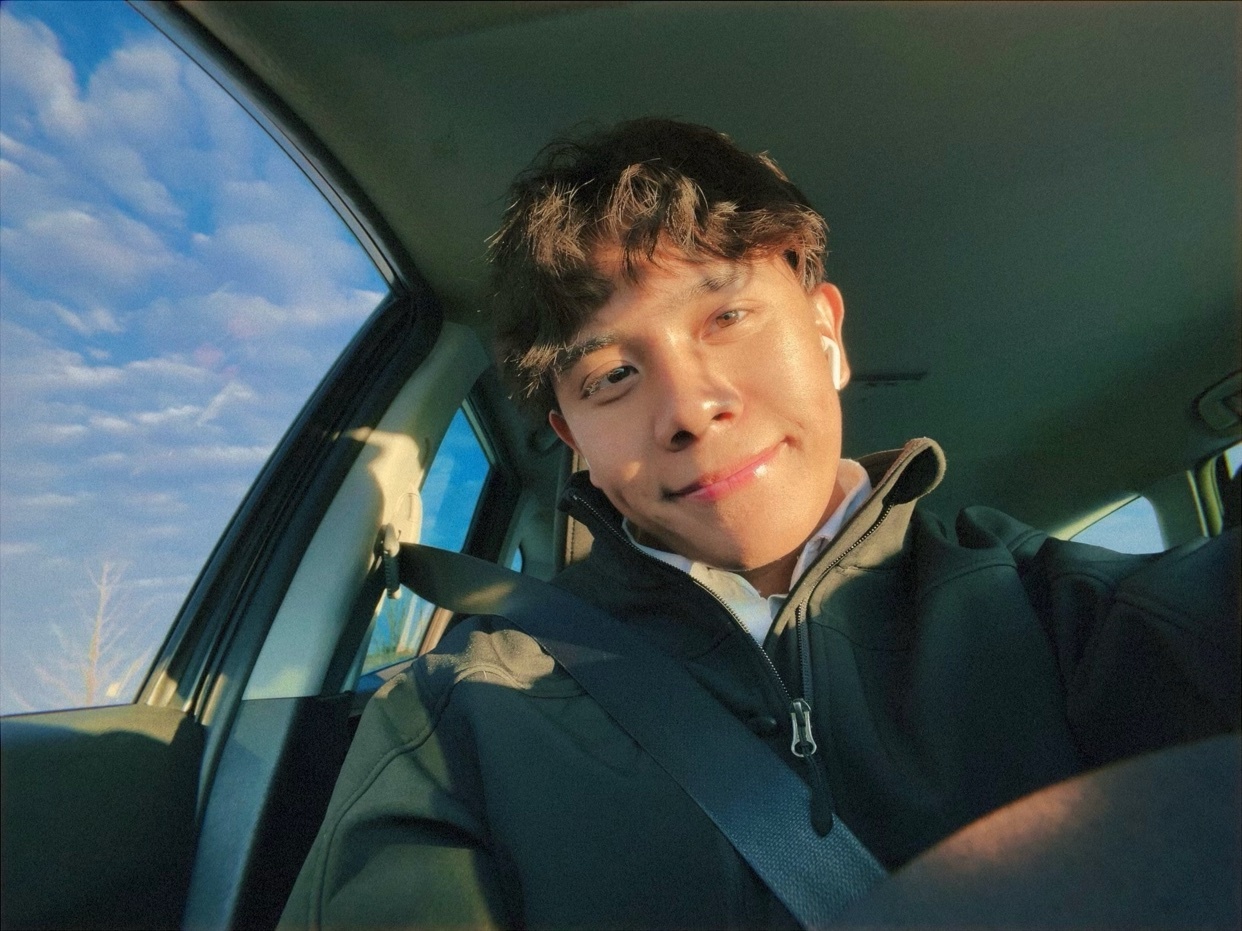 A young man smiles at the camera while sitting in the driver's seat of a car. He is wearing a dark jacket over a collared shirt and has earbuds in.