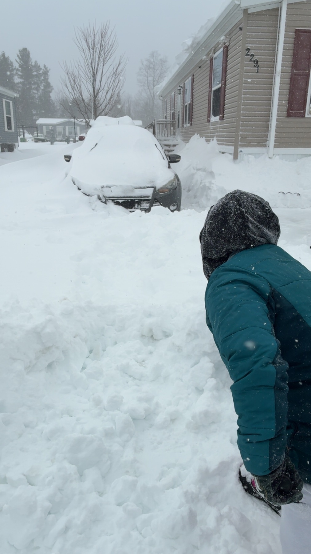 A person in a teal jacket and black gloves is partially visible in the foreground, facing away from the camera and towards a car completely buried in snow. The scene depicts a heavy snowfall with snowdrifts piled high around the vehicle and the surro