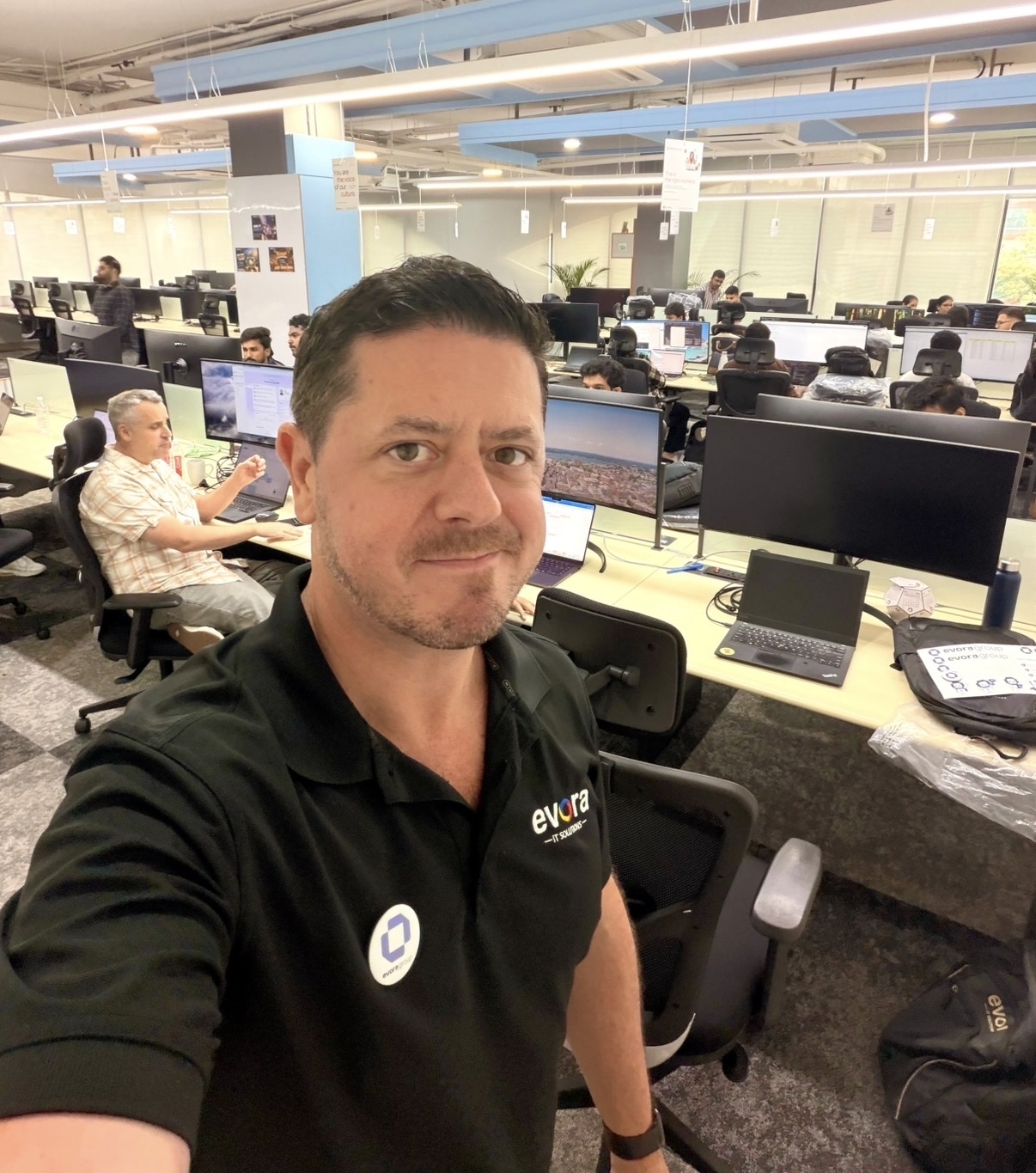 A man in a black polo shirt takes a selfie in a busy office environment. Rows of desks with computers and employees working fill the background.
