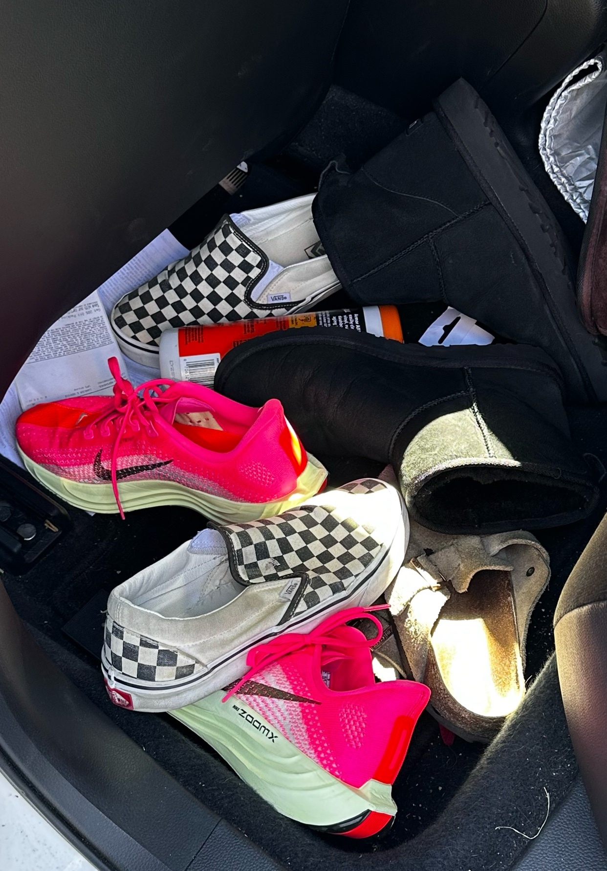 A pile of various shoes, including bright pink Nike running shoes, black Ugg boots, and checkered Vans, are jumbled together in the back of a car.