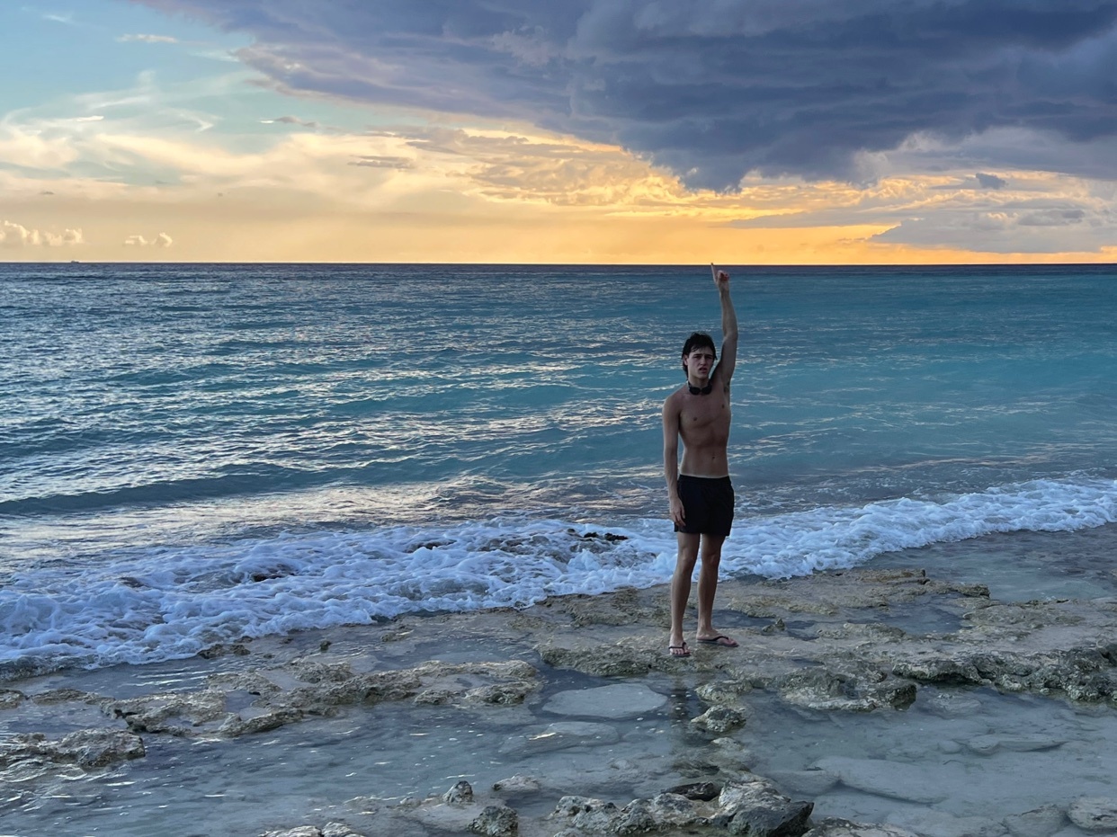A man stands on a rocky beach, raising his arm towards the sky. The photo captures a beautiful sunset over the ocean.