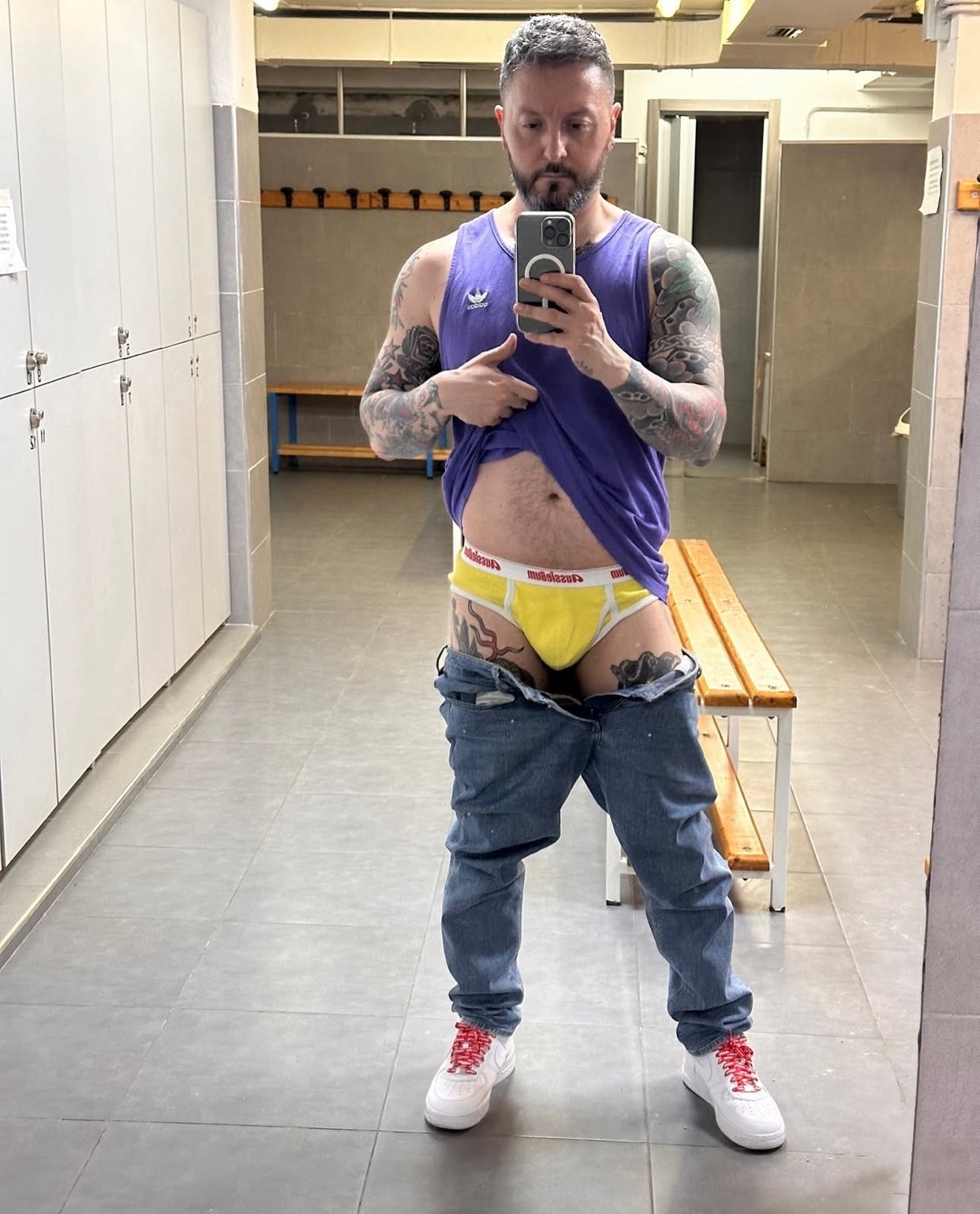 A man takes a mirror selfie in a locker room, showing off his physique and tattoos. He is wearing a tank top, jeans, and underwear.