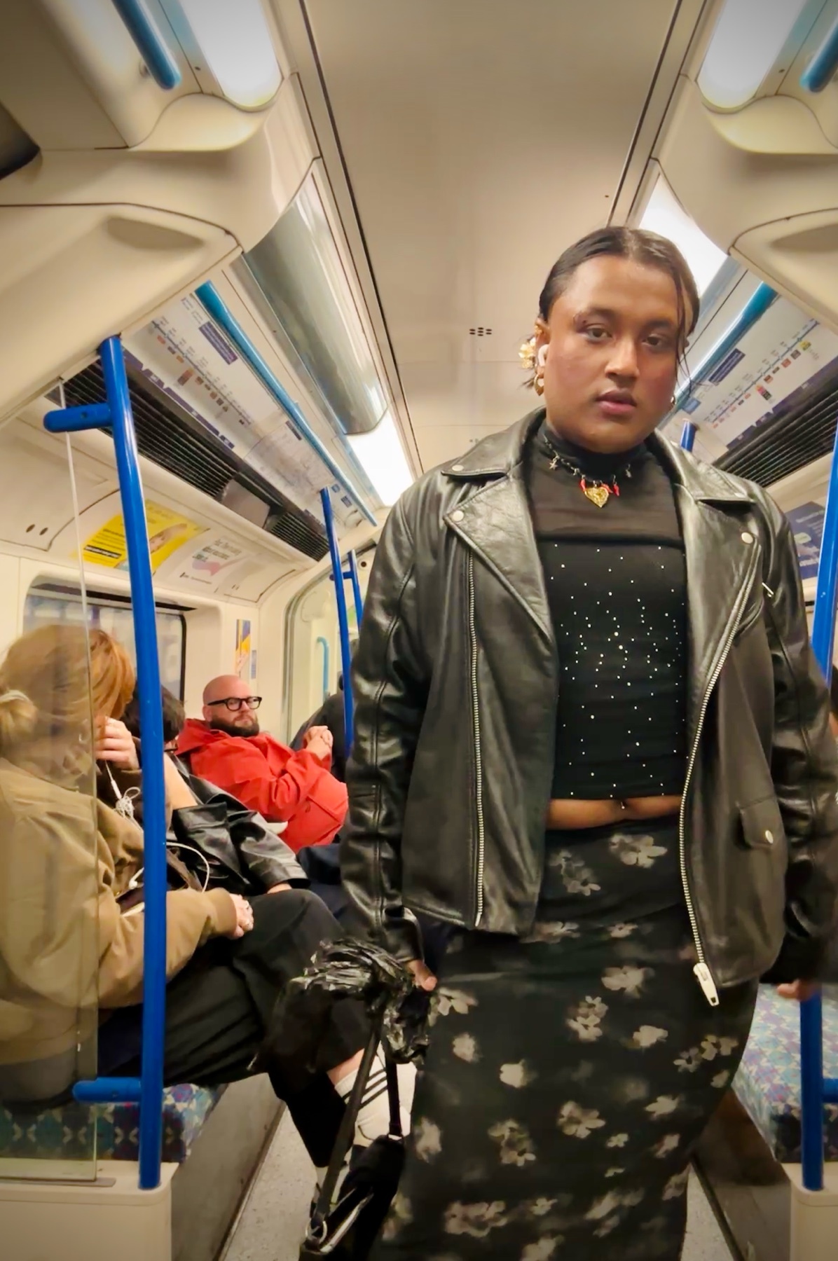 A non-binary person in a leather jacket and floral skirt stands on a train, looking towards the camera. Other passengers are visible in the background.