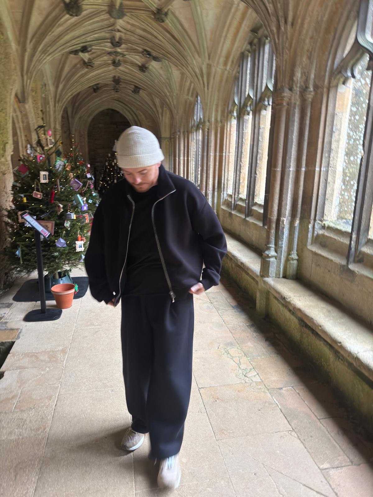 A man in a black outfit and a beanie stands in a hallway with Christmas trees. The architecture of the hallway is visible.