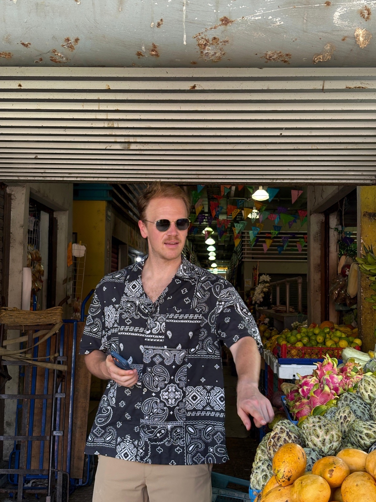 A man wearing sunglasses and a bandana print shirt stands in front of a fruit stand at an outdoor market. He is holding a phone in his left hand and reaching towards a display of exotic fruits.