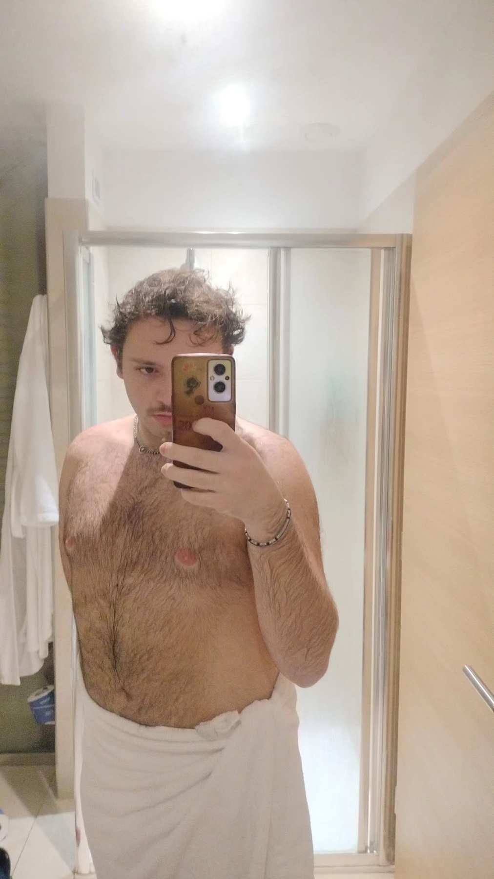 A man with curly hair and a hairy chest takes a selfie in a bathroom, wrapped in a white towel.