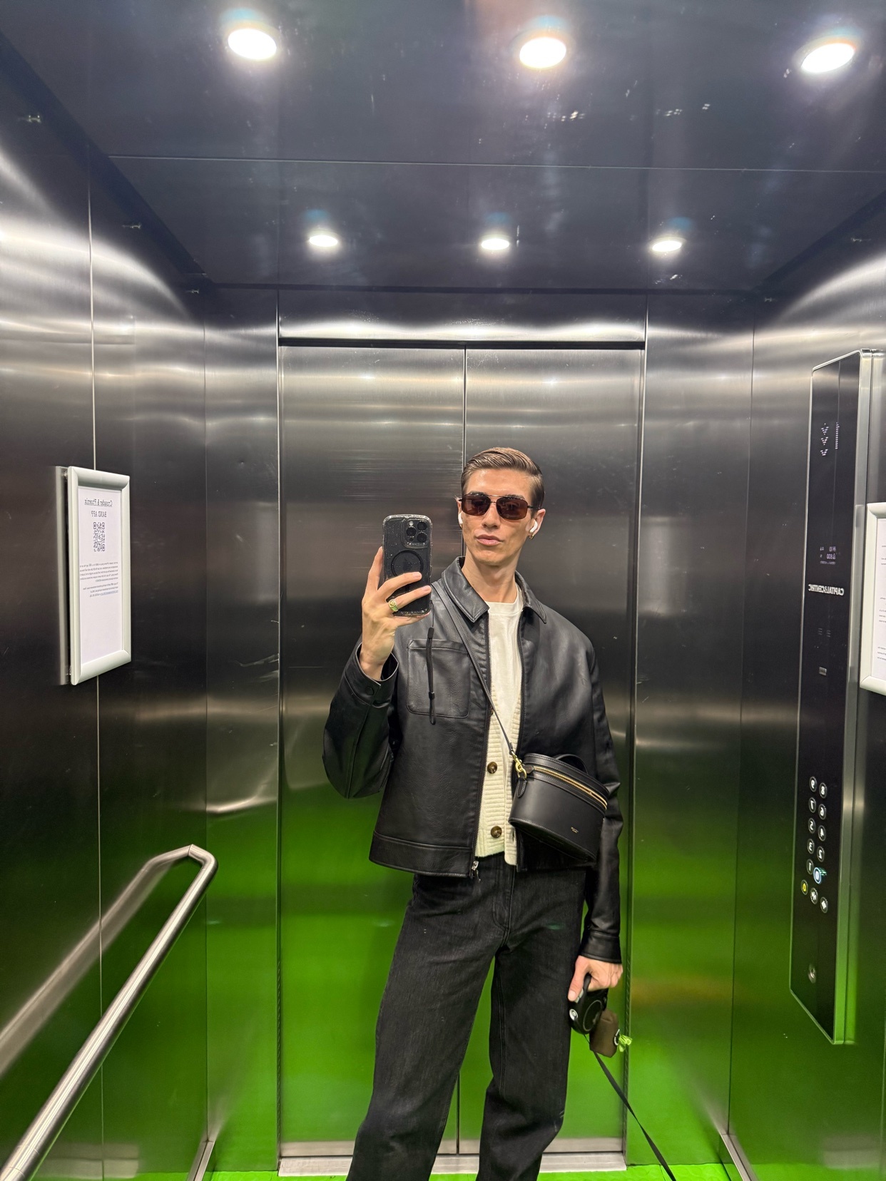 A man takes a mirror selfie in an elevator. He is wearing a black leather jacket, sunglasses, and jeans.