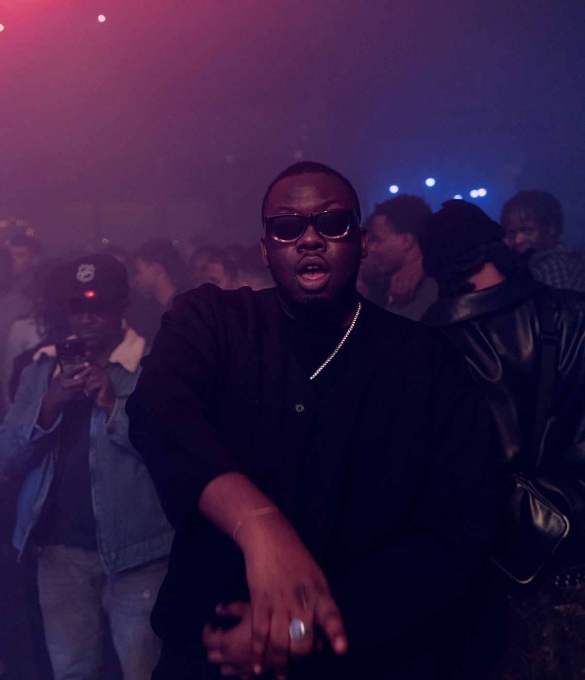 A man wearing sunglasses and a black turtleneck stands in a dimly lit, crowded venue. The atmosphere is energetic, with hints of blue and red lights illuminating the background.