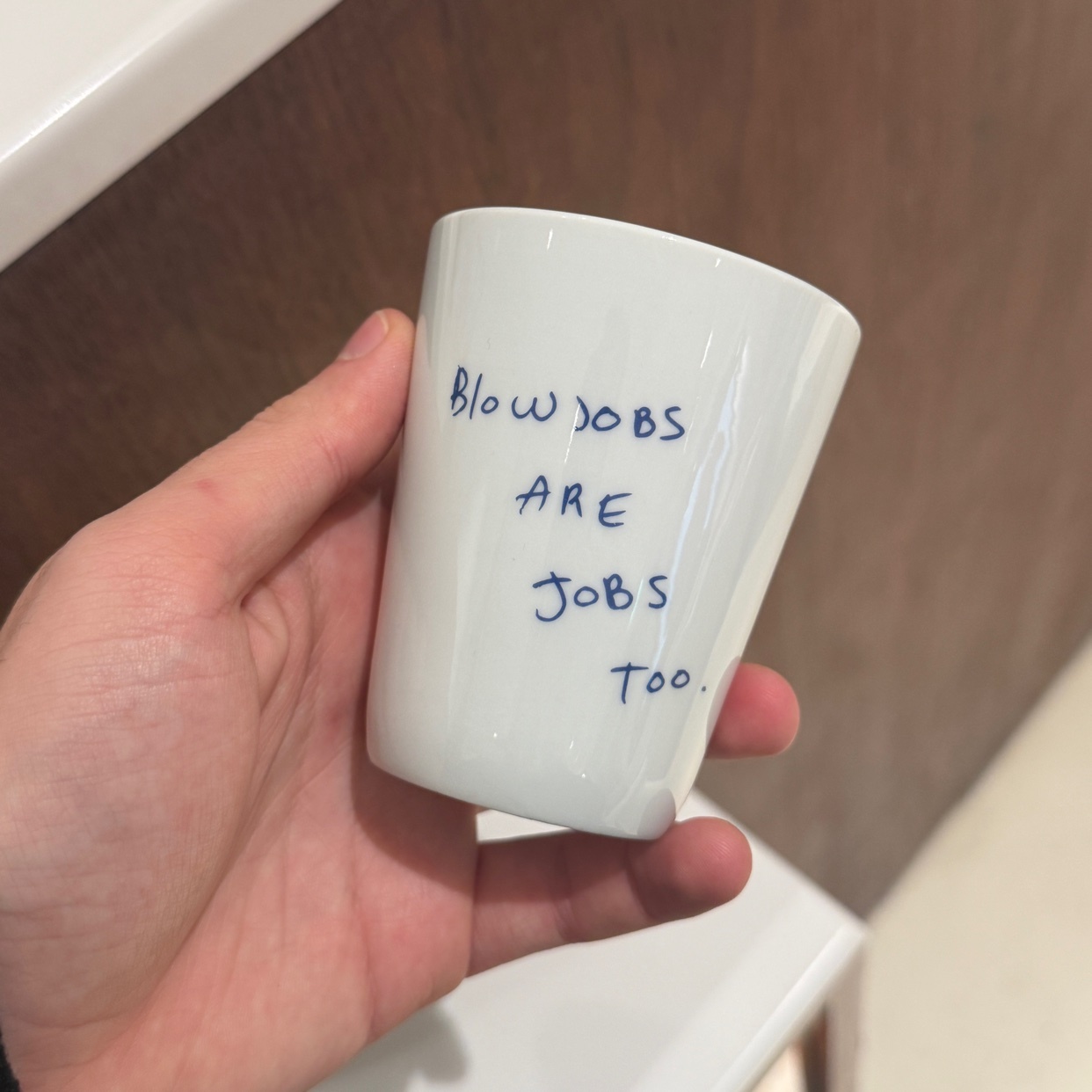 A hand holds a white cup with the words "Blow Jobs Are Jobs Too" written on it. The image is taken from a close-up perspective.