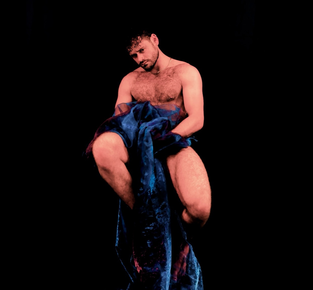 A shirtless man with dark hair and stubble poses against a black background, draped in blue fabric.