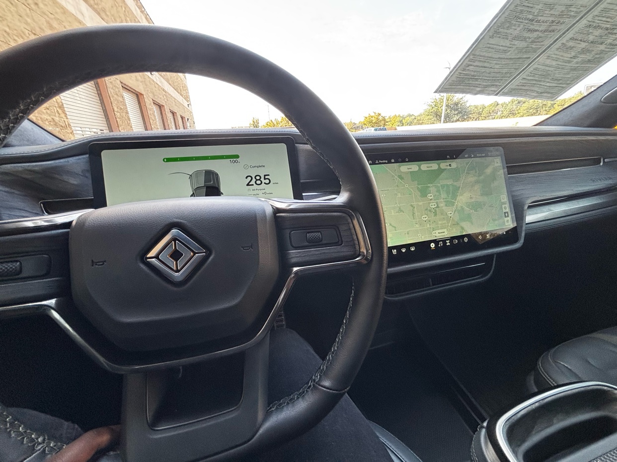 An interior shot of a Rivian electric vehicle. The dashboard features two large screens displaying navigation and vehicle information.