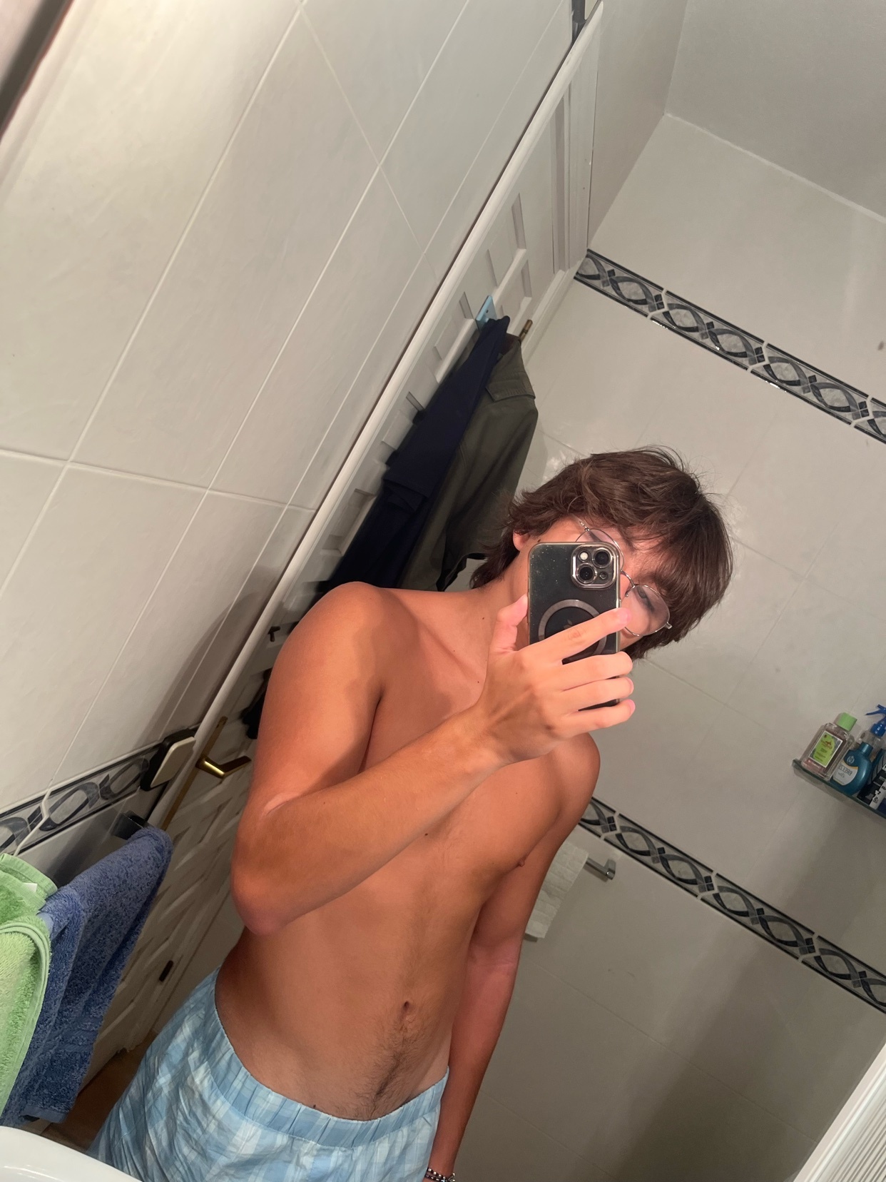 A shirtless man with tan lines takes a selfie in a bathroom mirror while wearing sunglasses.