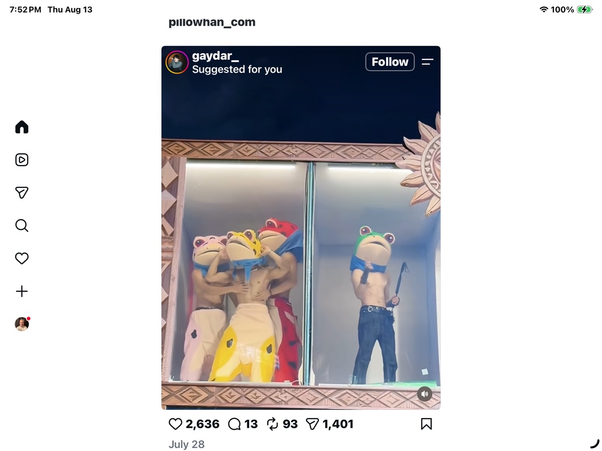 A group of people in frog costumes are displayed in a window. One person in the front right is shirtless, wearing jeans and a frog mask, holding a stick.