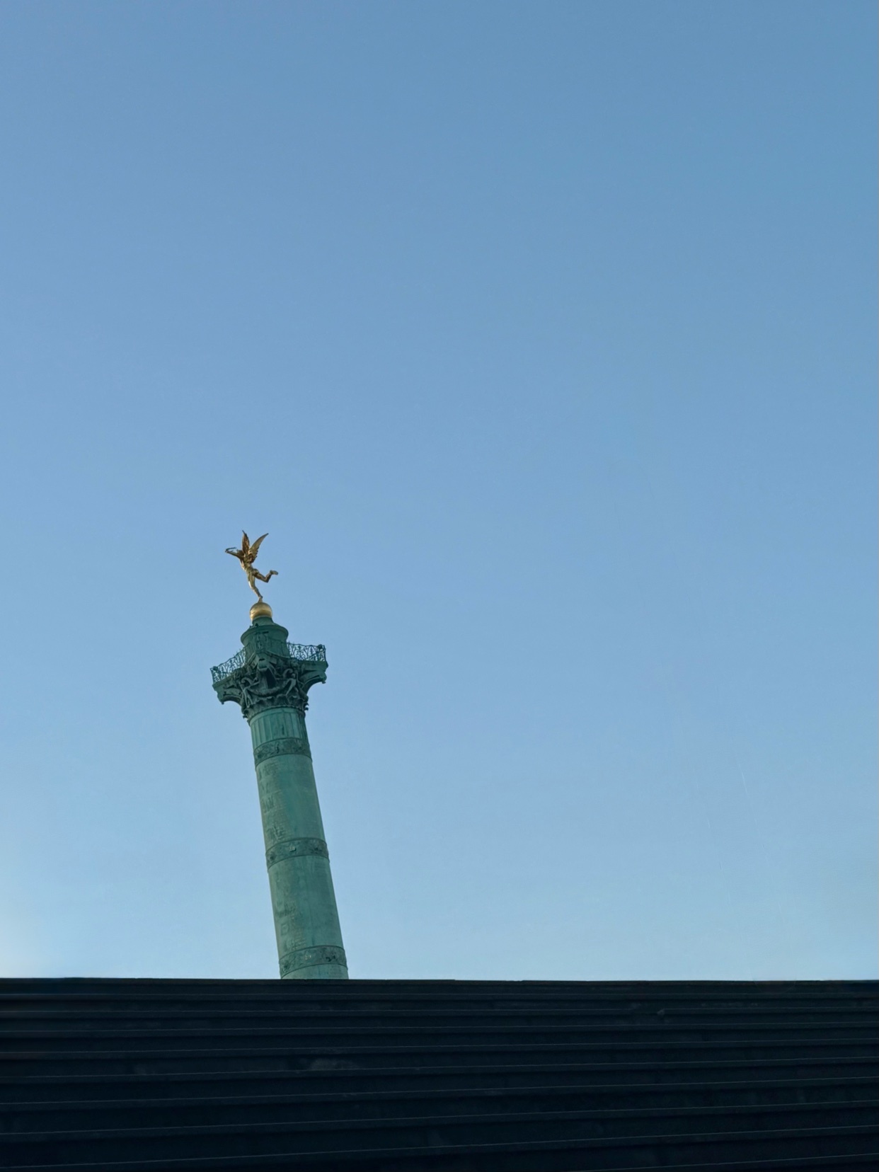 A golden angel statue stands atop a tall, green column against a clear blue sky. The base of the column is visible behind a dark, textured foreground.