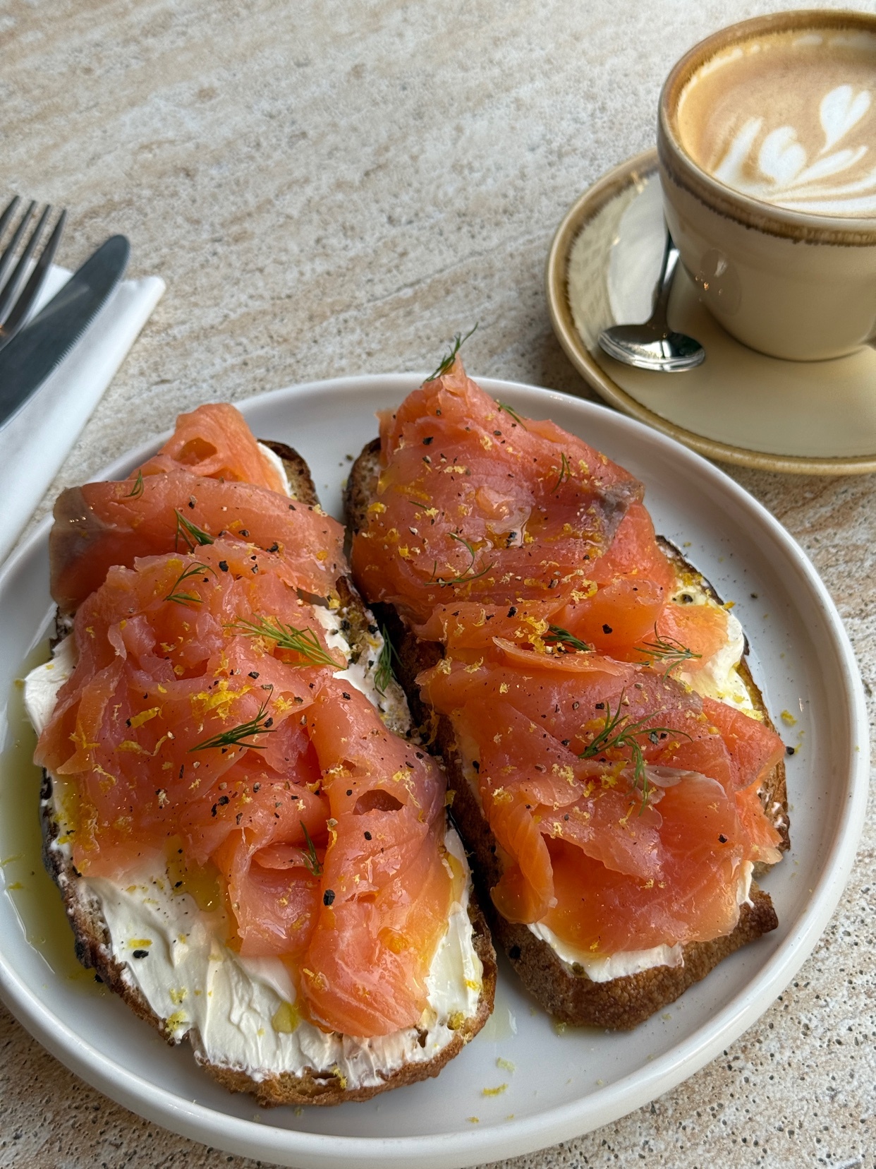 A close-up shot of two slices of toast topped with smoked salmon, cream cheese, and dill. A cup of coffee with latte art sits in the background.