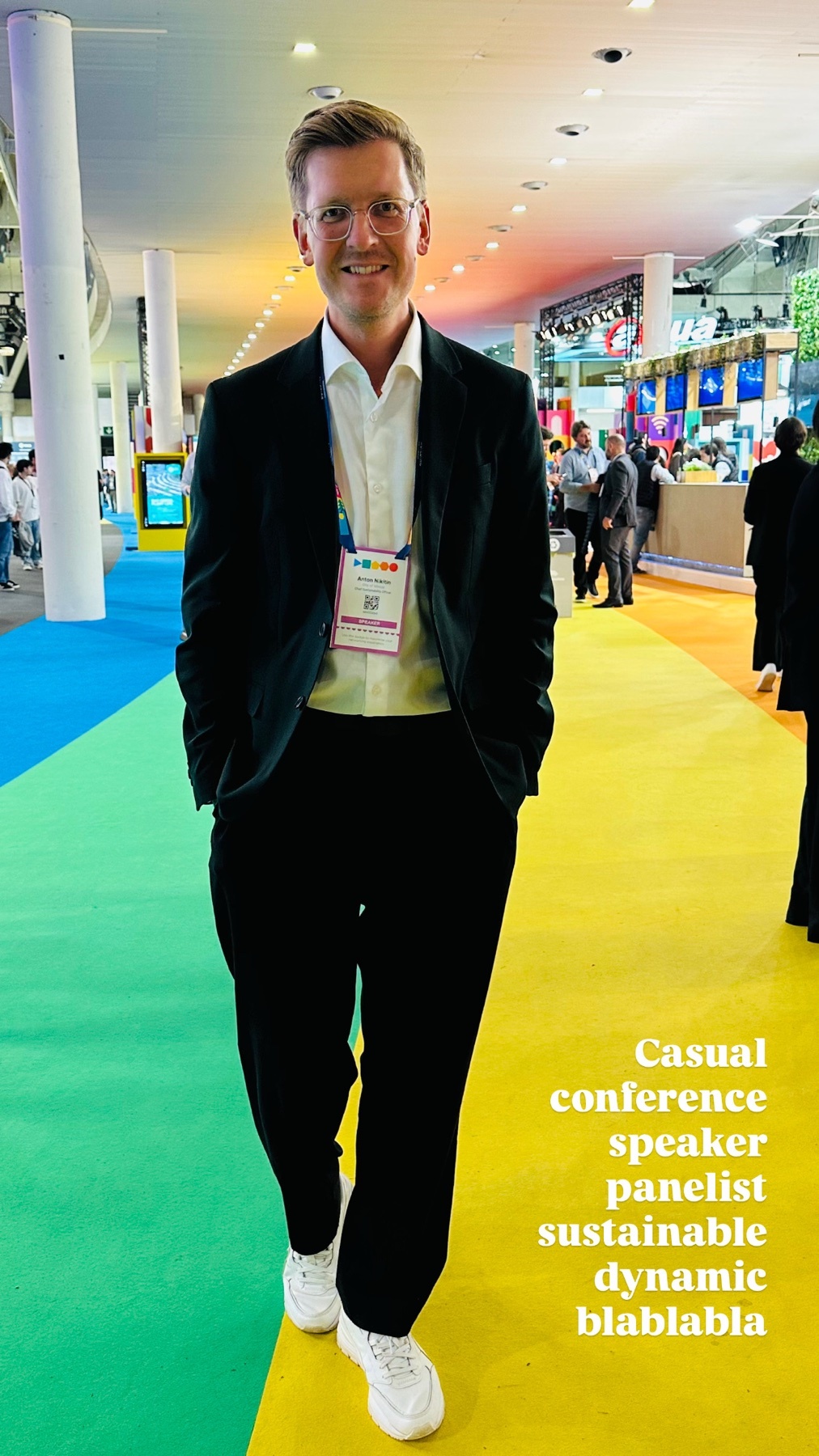 A man in a suit and sneakers walks towards the camera. He is smiling and wearing glasses.