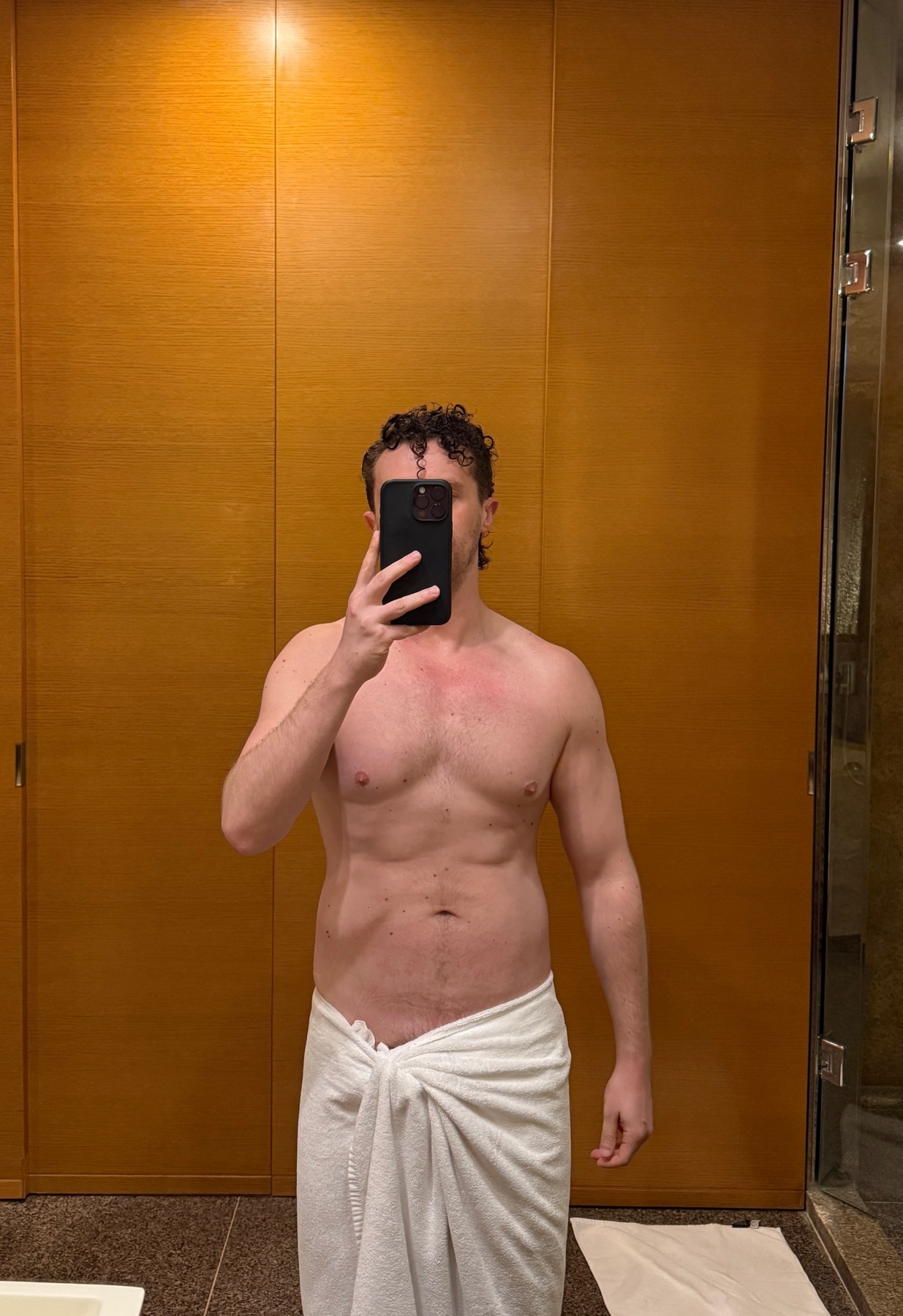 A man stands shirtless, wrapped in a white towel, taking a mirror selfie in a bathroom. The warm wooden walls and tiled floor suggest a spa-like or hotel setting.