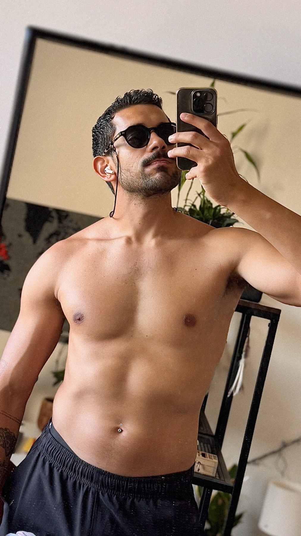 A man with sunglasses and stubble takes a mirror selfie, showing off his muscular physique and a new belly button piercing.
